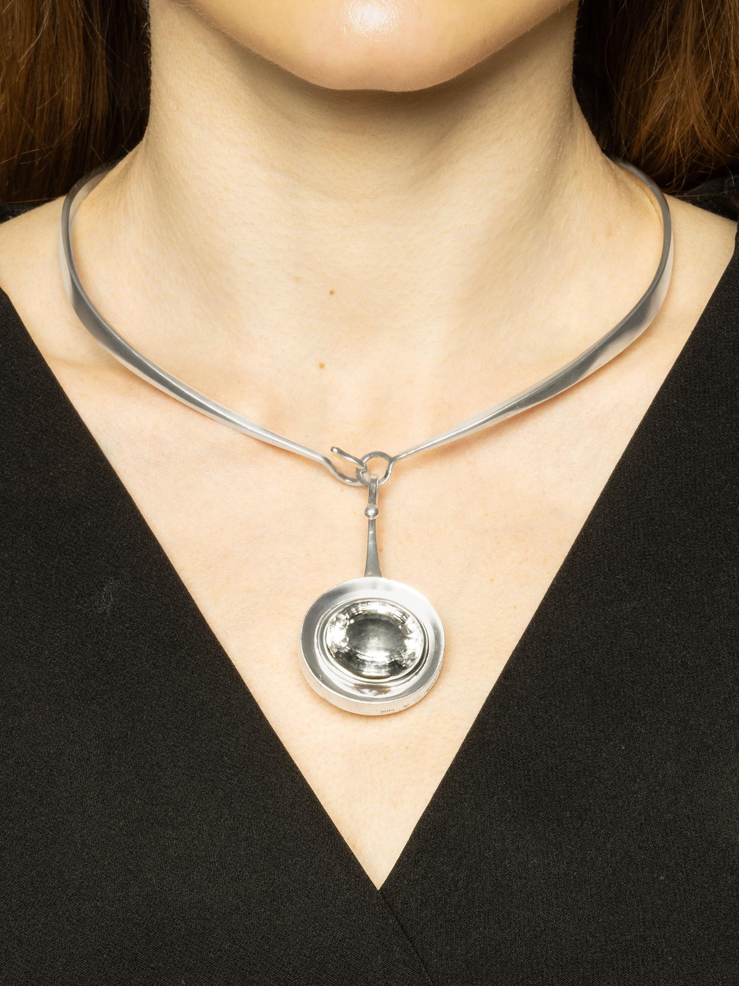 This sterling silver neck ring features a unique twisted design, with its shape going back and forth rather than tapering gradually towards the back. The pendant, with its hook and oval shape, showcases an oval faceted rock crystal that takes on an