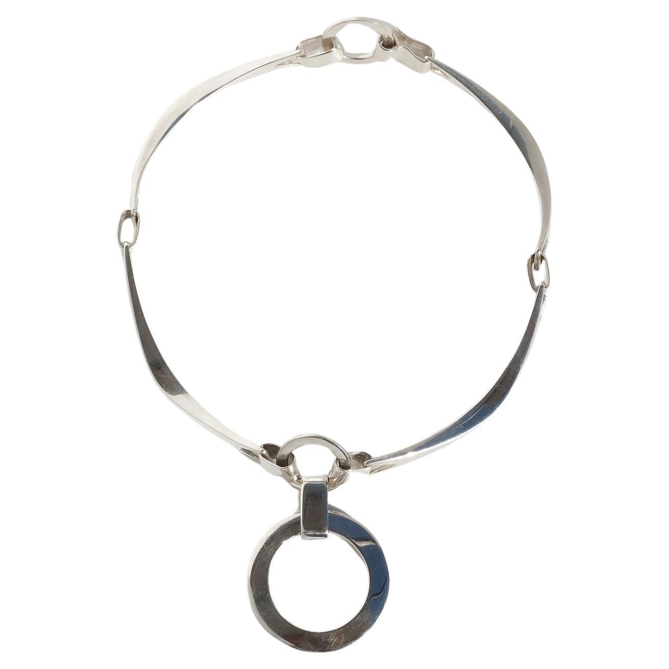 This sterling silver necklace is adorned with a sterling silver circle pendant. The necklace itself consists of four arched silver bars which are attached to each other with five silver links and two smaller silver circles. The necklace opens and