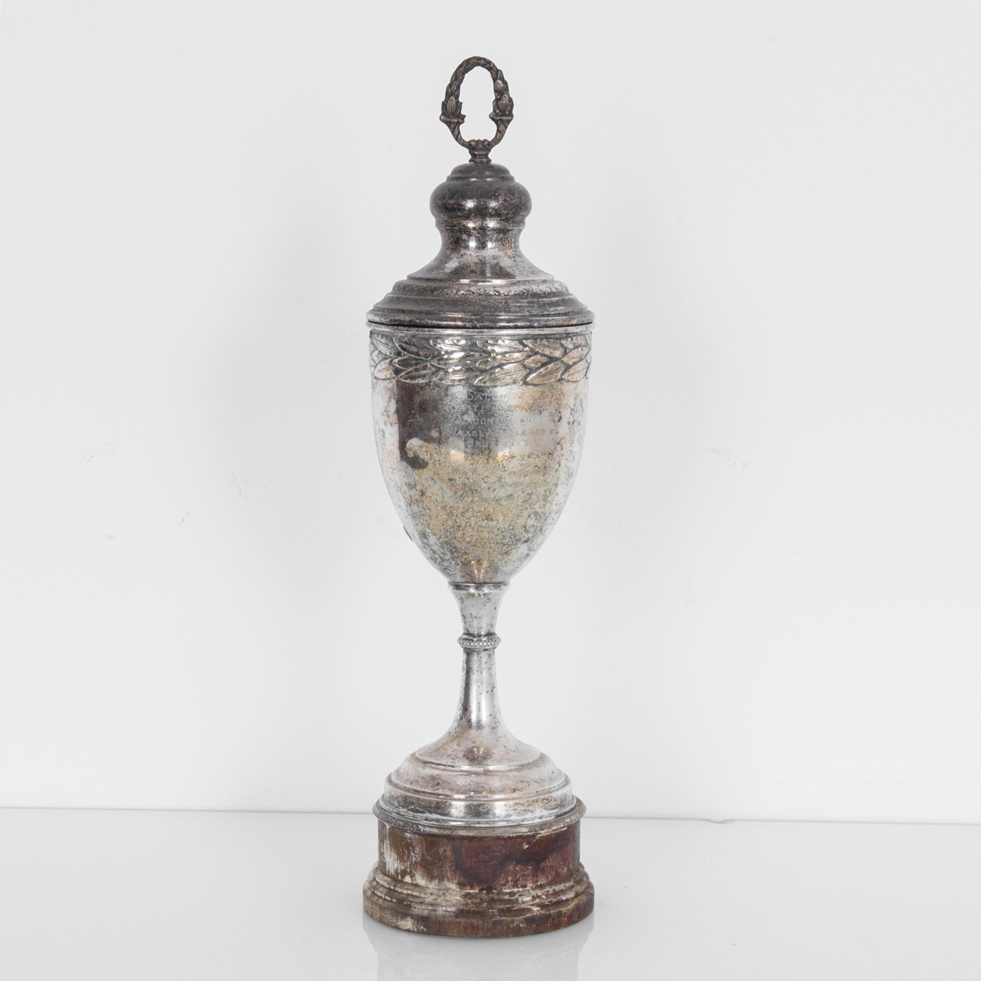 A silver plated trophy from Europe, made circa 1900-1930. A slender silver cup topped with a victory wreath, girdled with a circle of laurel leaves. The tarnishing of the silver obscures the words engraved into the surface, lending the trophy a