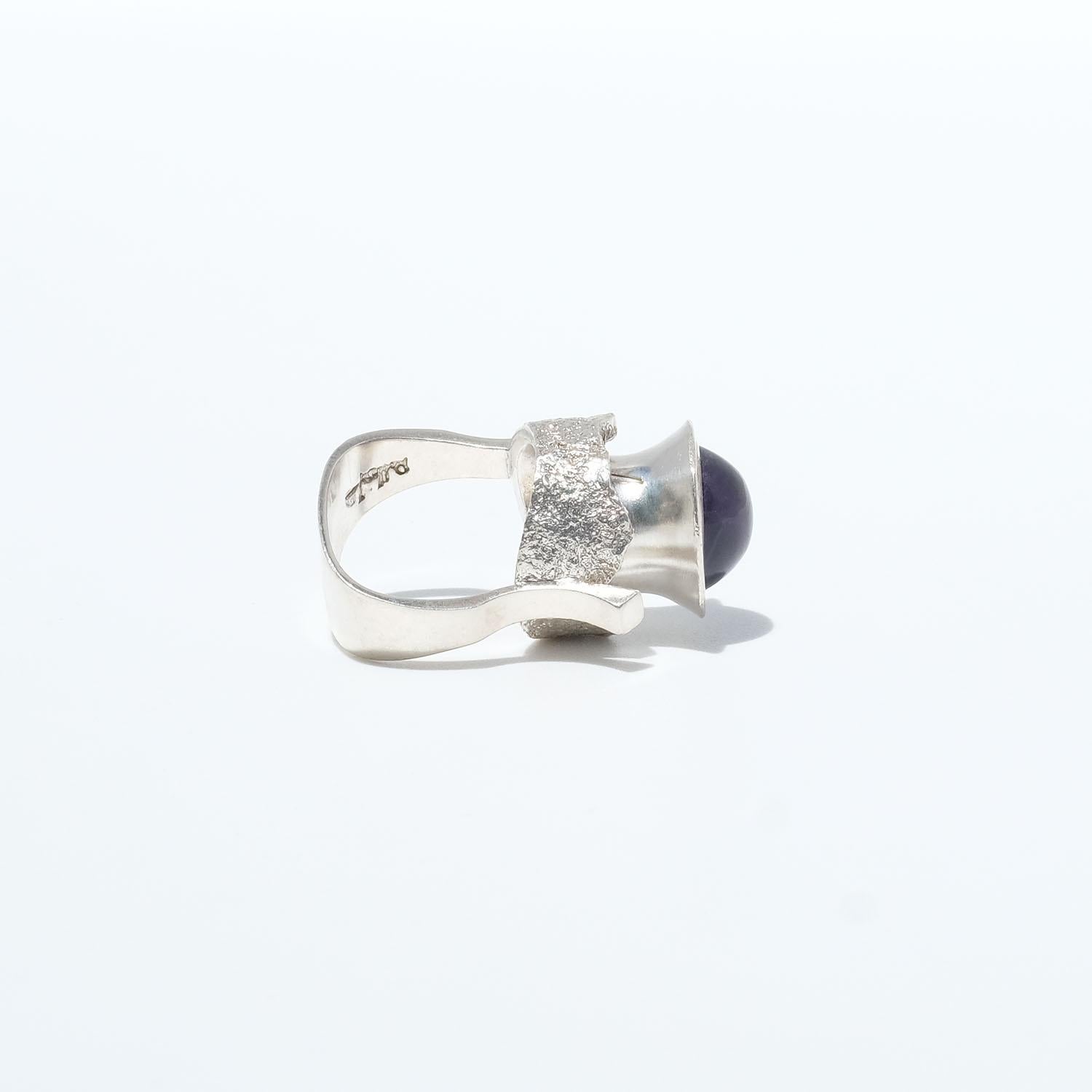 Este anillo modernista de plata está adornado con una amatista tallada en cabujón que está montada sobre algo que parece un barril de plata. Tanto el engaste como la caña del anillo son prominentes. La plata de la parte inferior del engaste, la más