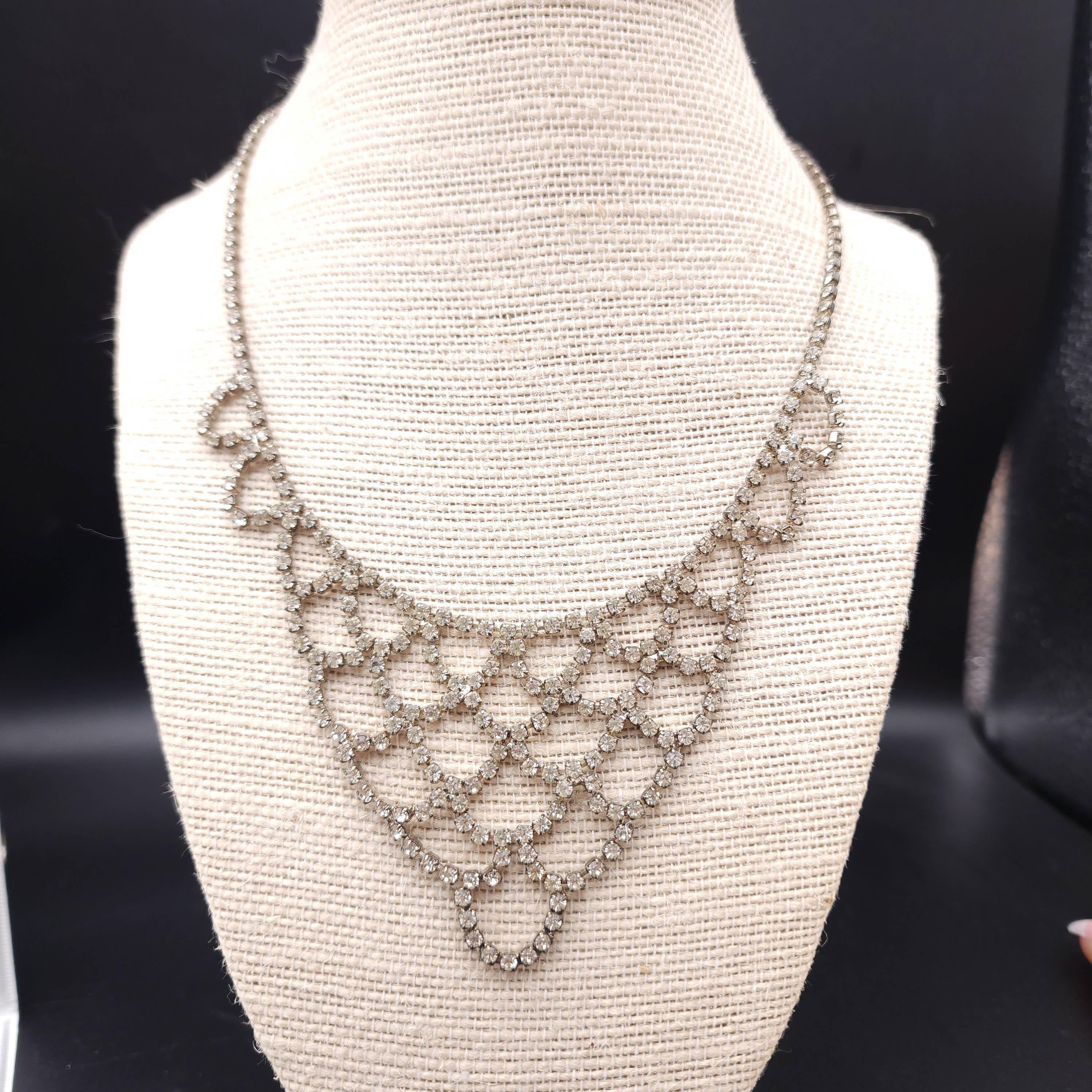 This stunning vintage bib necklace features a cascading, scalloped design adorned with sparkling rhinestones set in a silver-tone metal. 

The intricate latticework creates a glamorous statement piece that elegantly drapes over the neckline, perfect