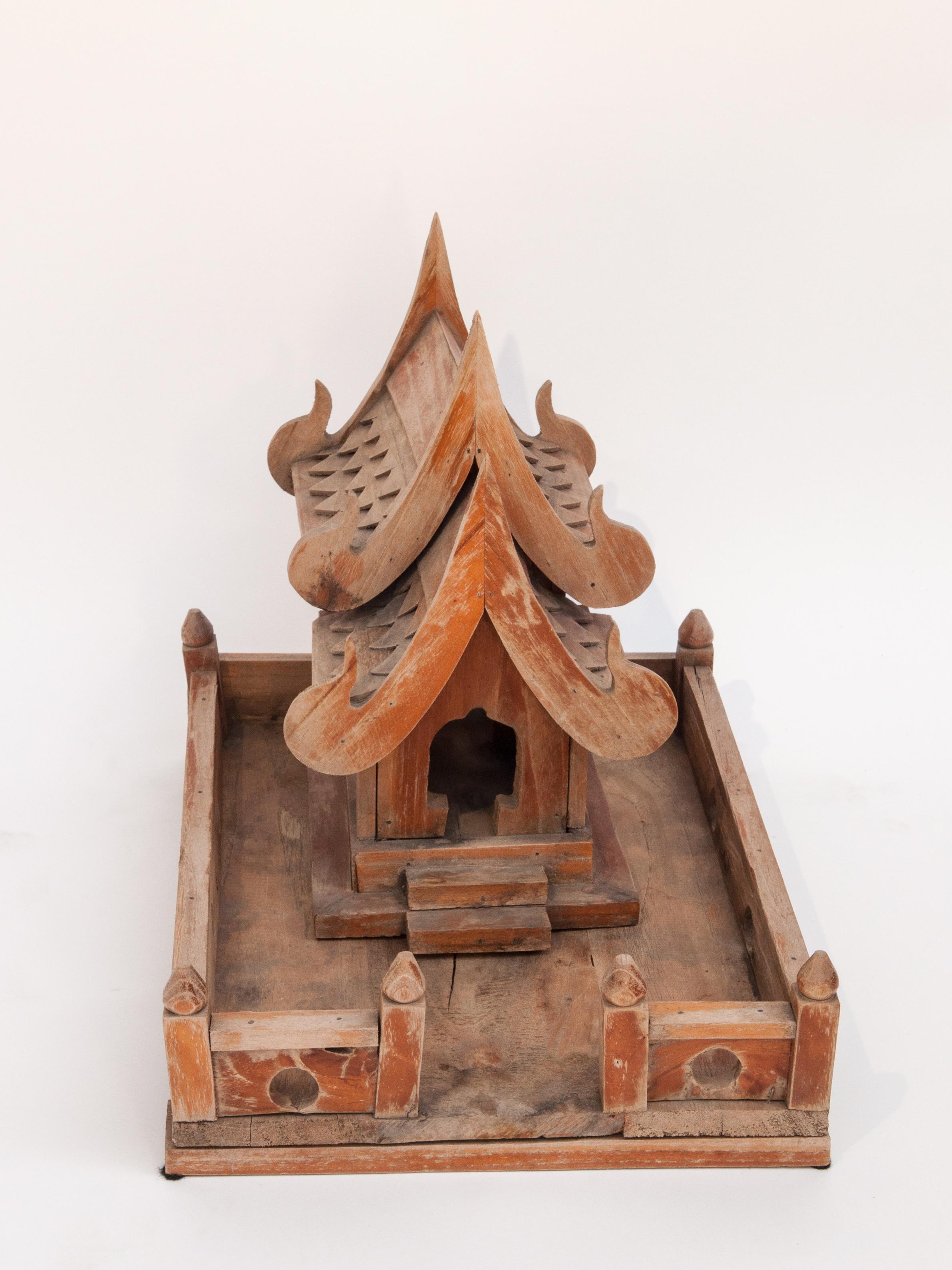 Vintage small spirit house from Northern Thailand. Teak, mid-late 20th century.
This simple rustic spirit house models a traditional Thai countryside temple, with fence and compound, and incorporates the Naga motif barge boards running along the