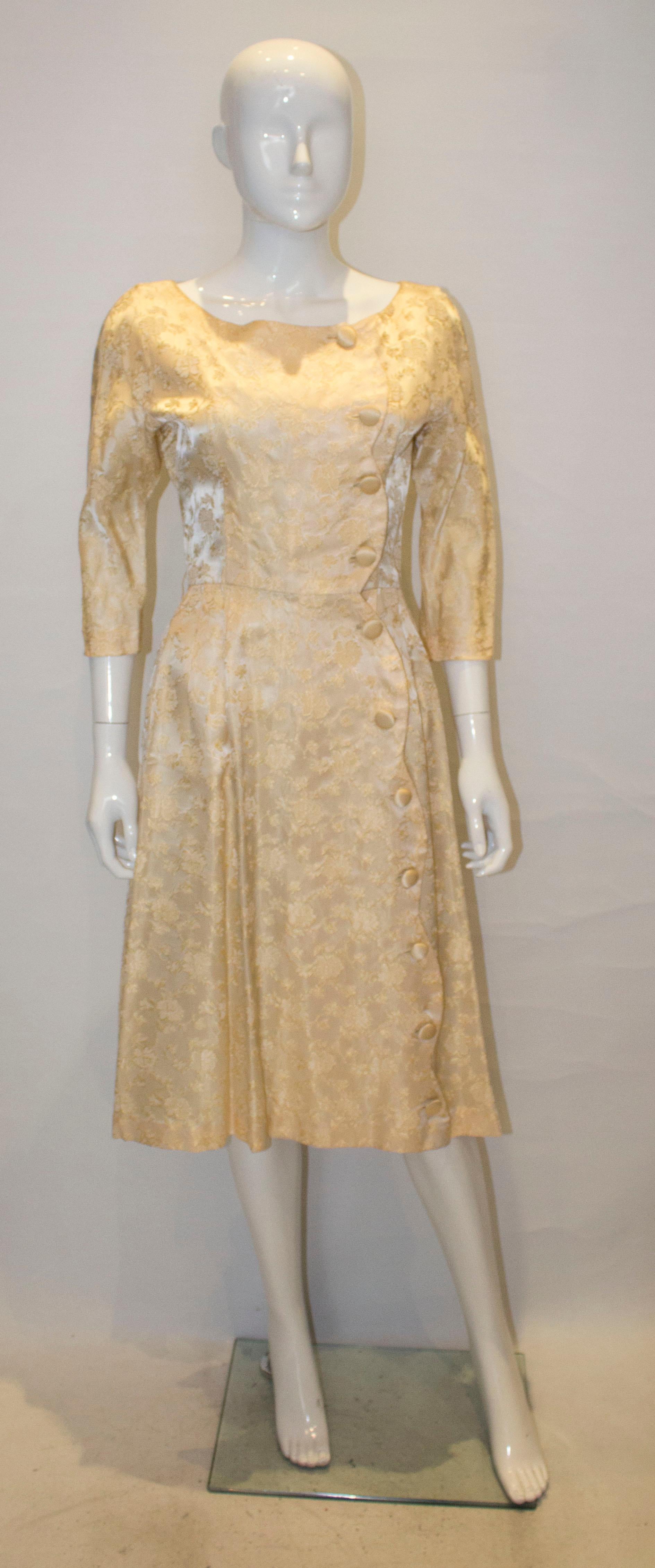 A pretty soft gold colour vintage brocade cocktail dress. The dress has a scallop edge opening with button fastenings. It has elbow length sleaves  and gathering at the back.
