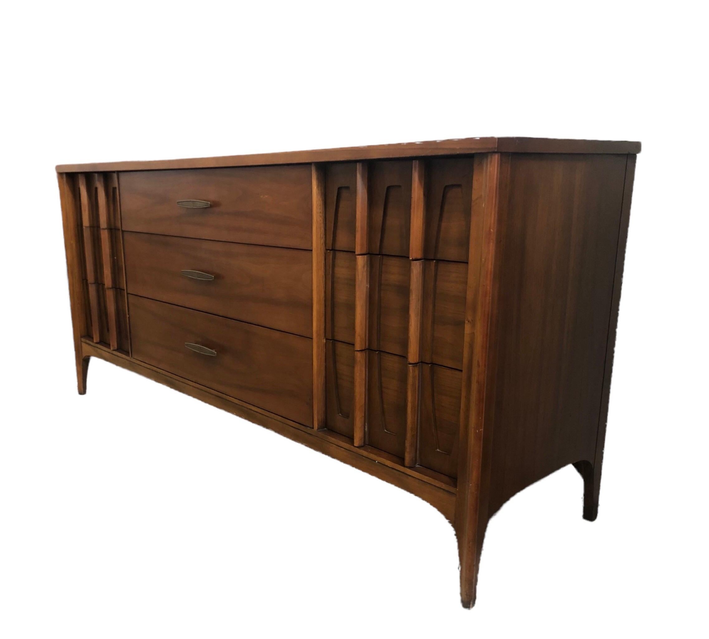 This stylish Kent Coffey triple dresser or credenza has lovely recessed scalloped accents and vertical wood pulls! Sleek and sculpted design with textured brass pull down the three middle drawers and solid walnut wood.

Measures: 64