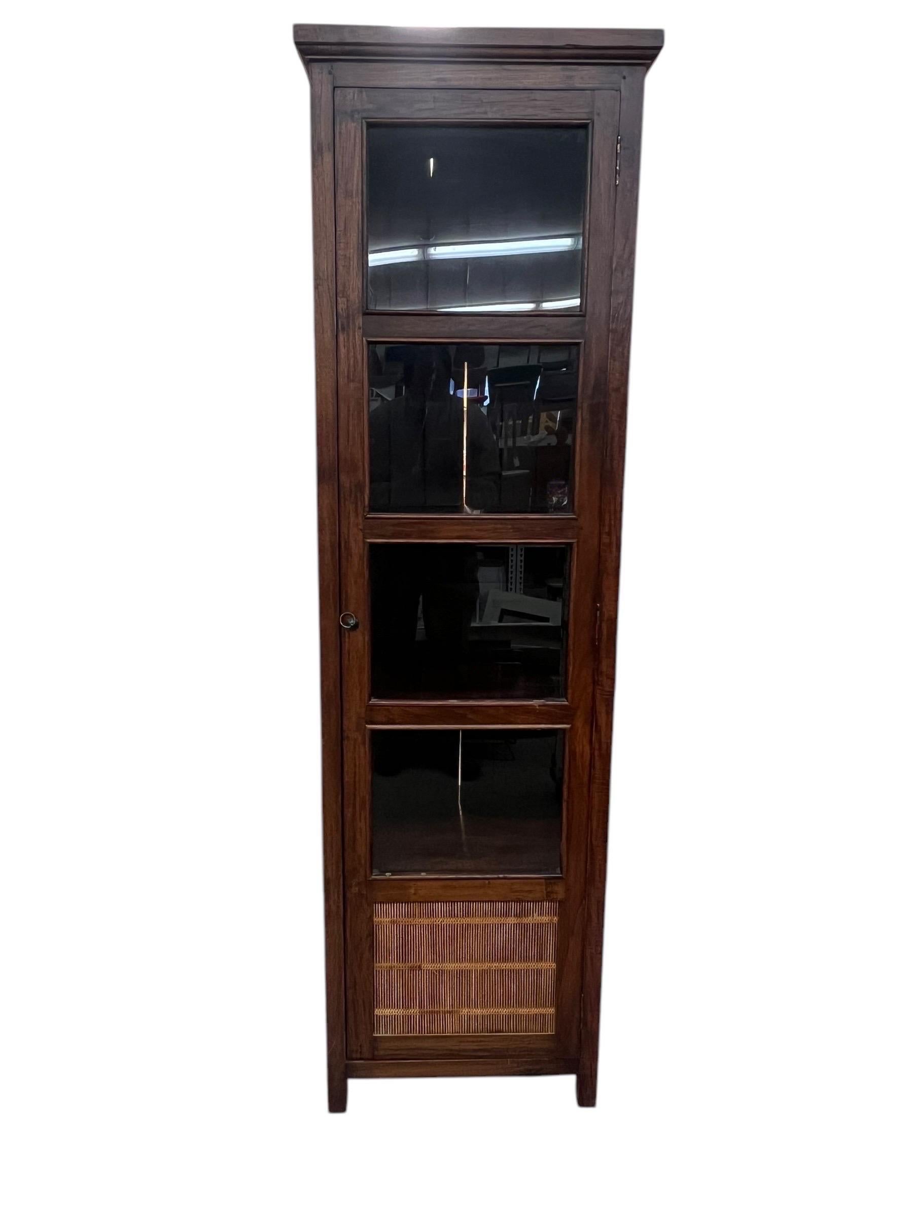 This vintage cabinet showcases a tall, narrow design, featuring rattan details on its sides and glass panels on the doors. The combination of wood and rattan gives it a distinctive, tropical aesthetic. The cabinet includes multiple shelves, offering