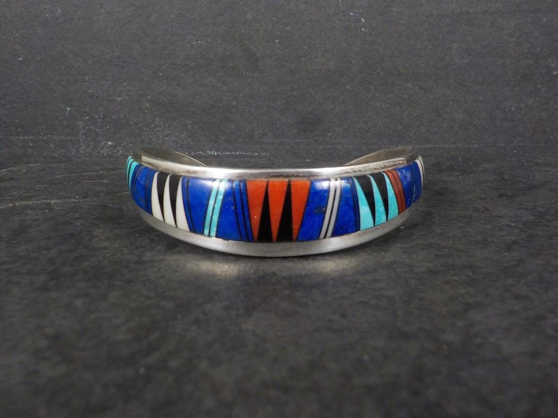 This gorgeous cuff bracelet is sterling silver.
It features inlay in lapis, jet, turquoise, mother of pearl, and coral.

The bracelet measures 5/8 of an inch at its widest and has an inner circumference of 6.25 inches, including the 1 1/4 inch