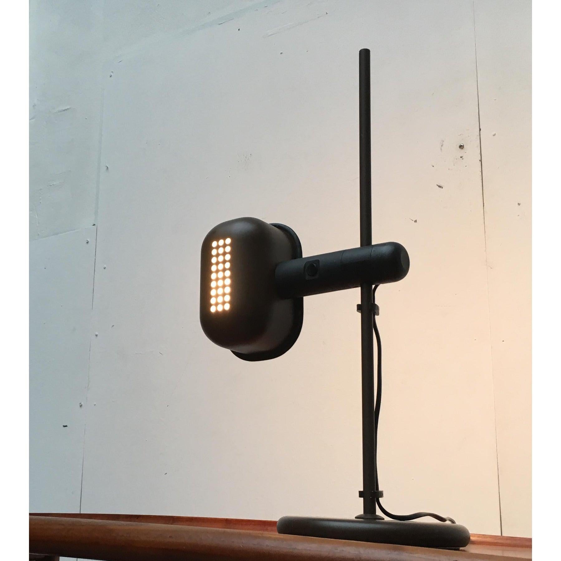Lampe de table NO AGE en brun et noir.

La lampe a un abat-jour rectangulaire avec des bords arrondis et une perforation distinctive sur le dessus. Le bord inférieur est fermé par un caoutchouc noir. La tête de la lampe est rotative et peut être