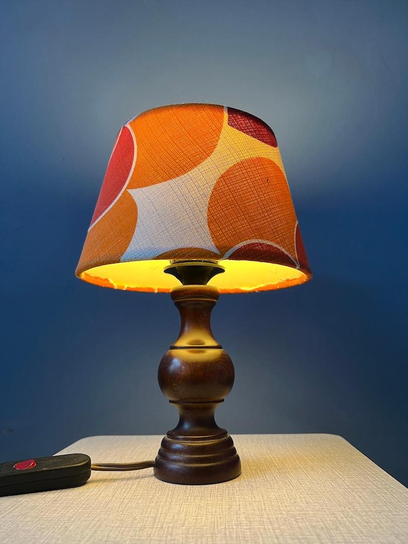 Small retro table lamp with newly made orange, space age style shade. The lamp has a wooden base and textile shade. The lamp requires one E27 lightbulb and currently has an EU-plug.

Additional information:
Materials: Ceramic, cotton
Period: