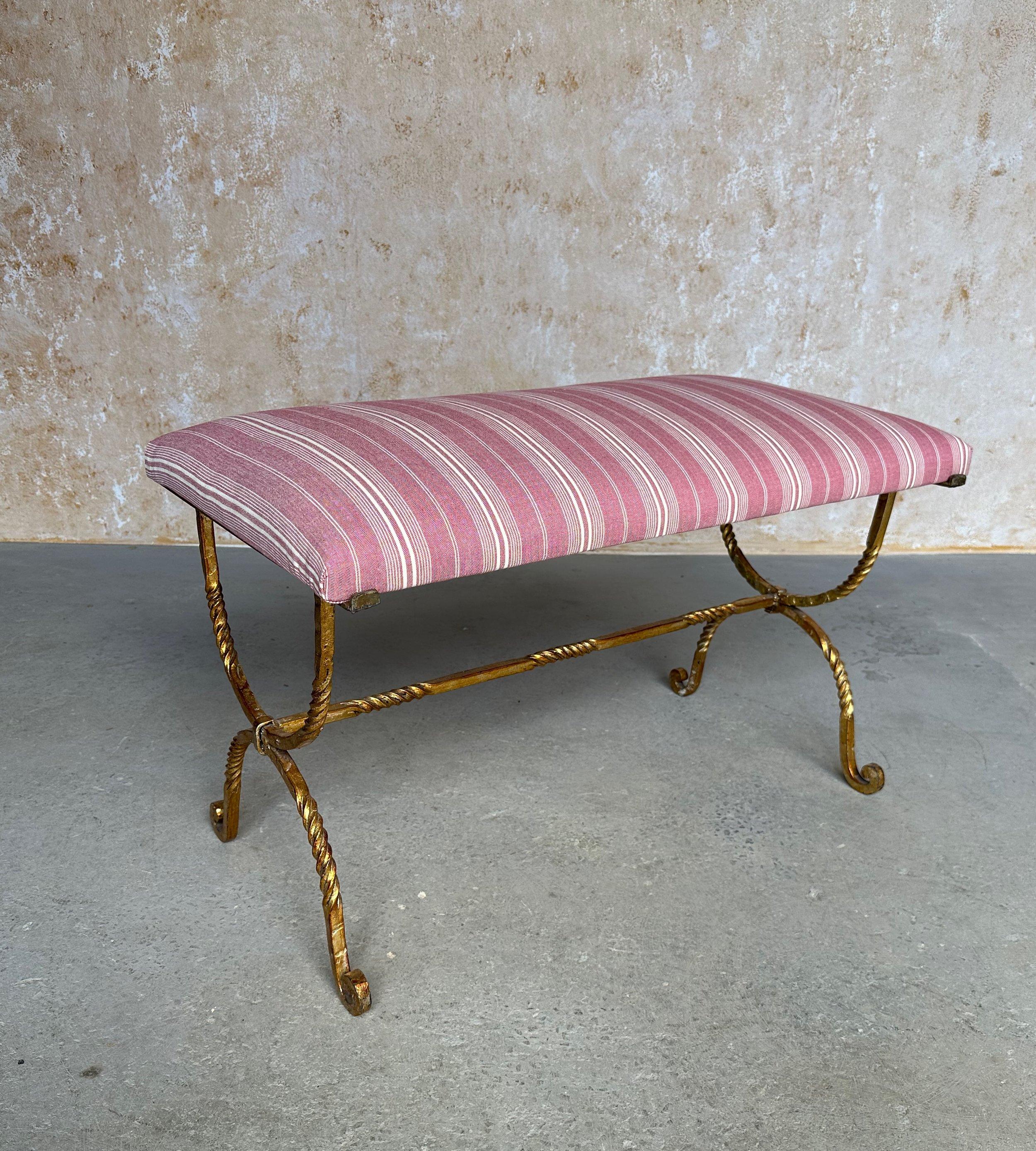 This lovely vintage 1950s Spanish gilt iron bench features an ornate hand forged twisted hourglass frame with central stretcher and beautifully scrolled feet. The rich gilt finish is hand-applied and the seat is upholstered in fashionable mauve