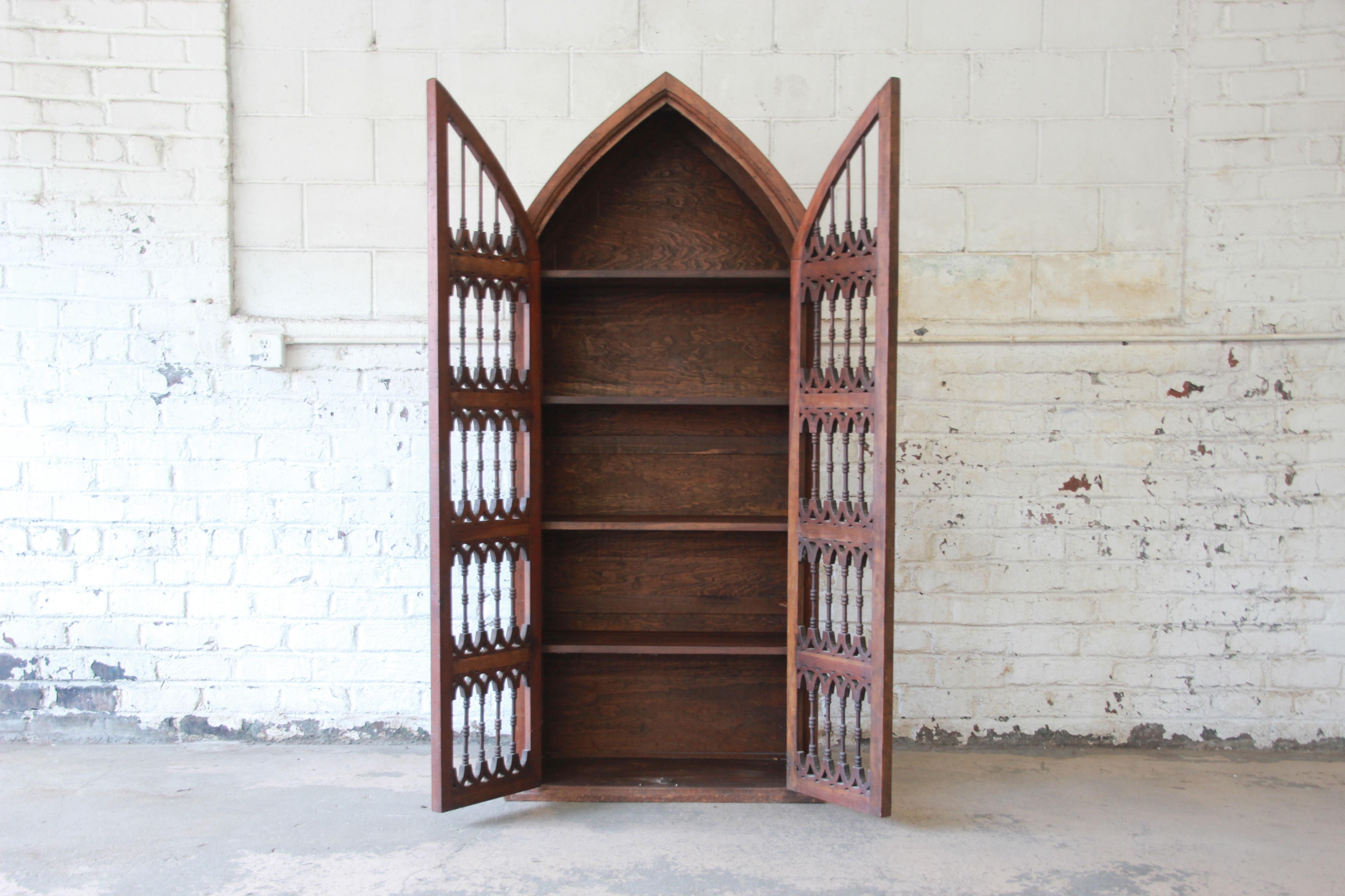 A very unique vintage Spanish Gothic Cathedral style bookcase or cabinet. The bookcase features solid wood construction with beautiful rustic dark pine wood grain and a unique cathedral shape. It has two doors with gorgeous carved spindles and