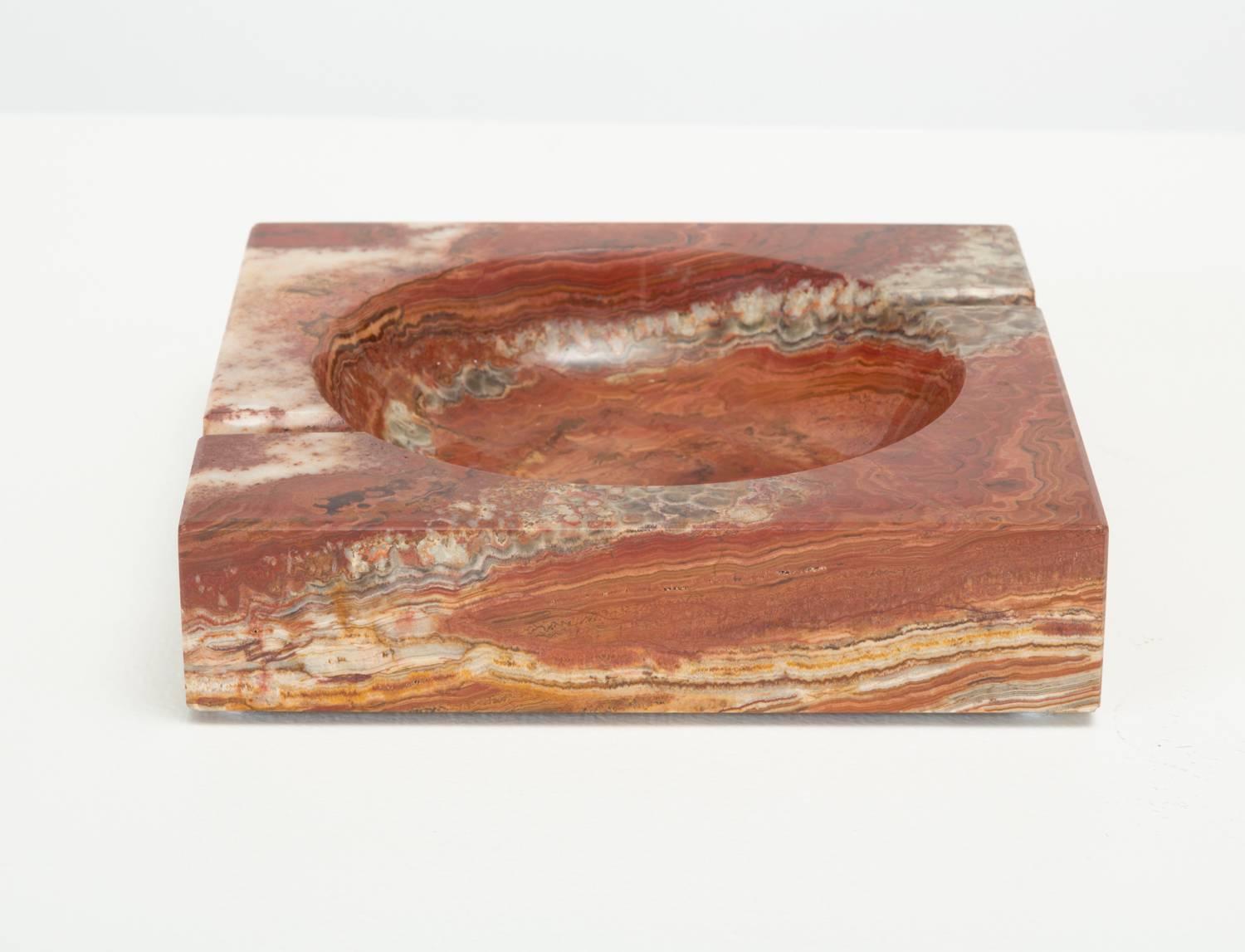 A square ashtray in a polished red marble with grey and white veining. The piece has a circular well, and two routed indentations perpendicular to its edges. The corners and edges of the face are chamfered and the piece has four felt feet on the
