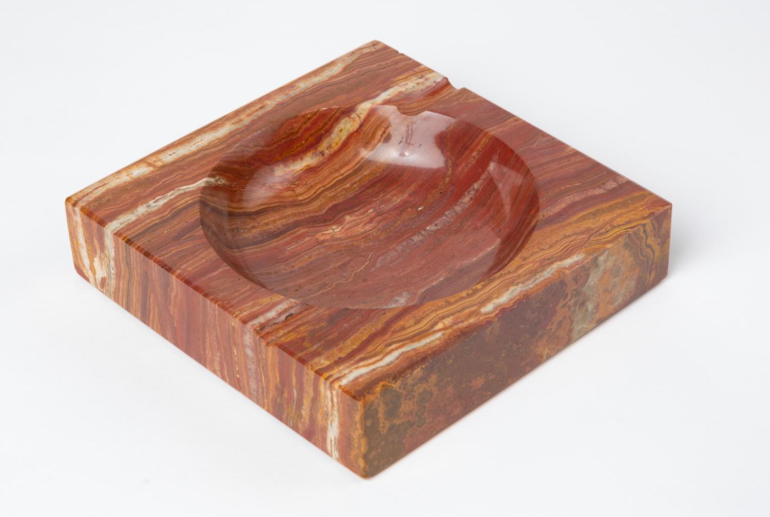 A square ashtray in a polished red marble with gray and white veining. The piece has a circular well, and two routed indentations perpendicular to its edges. The corners and edges of the face are chamfered and the piece has a red felt pad on the