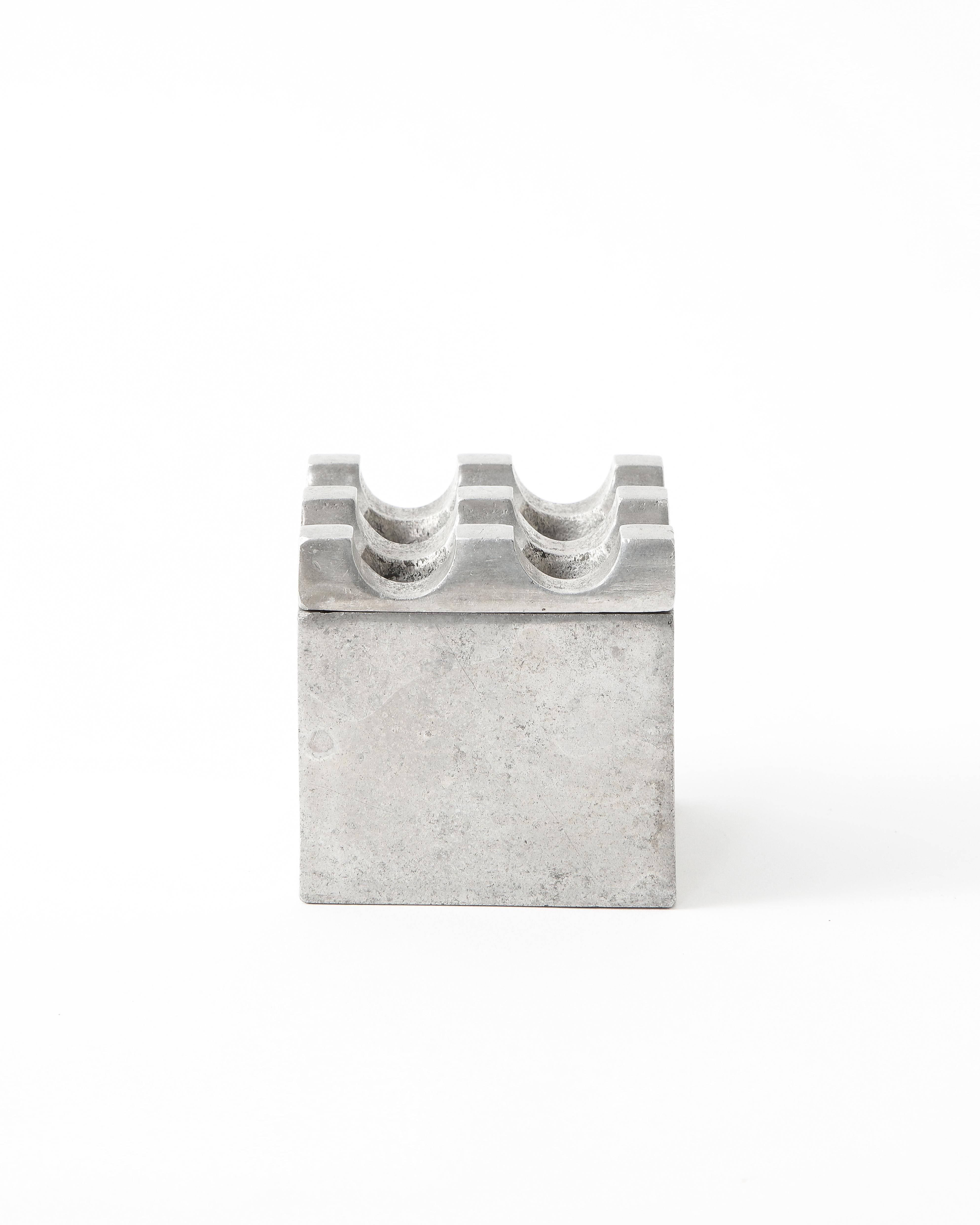 Pequeño cenicero vintage cuadrado de aluminio liso y muy desgastado. La parte superior extraíble presenta un dibujo cuadriculado con cuatro grandes perforaciones. 
