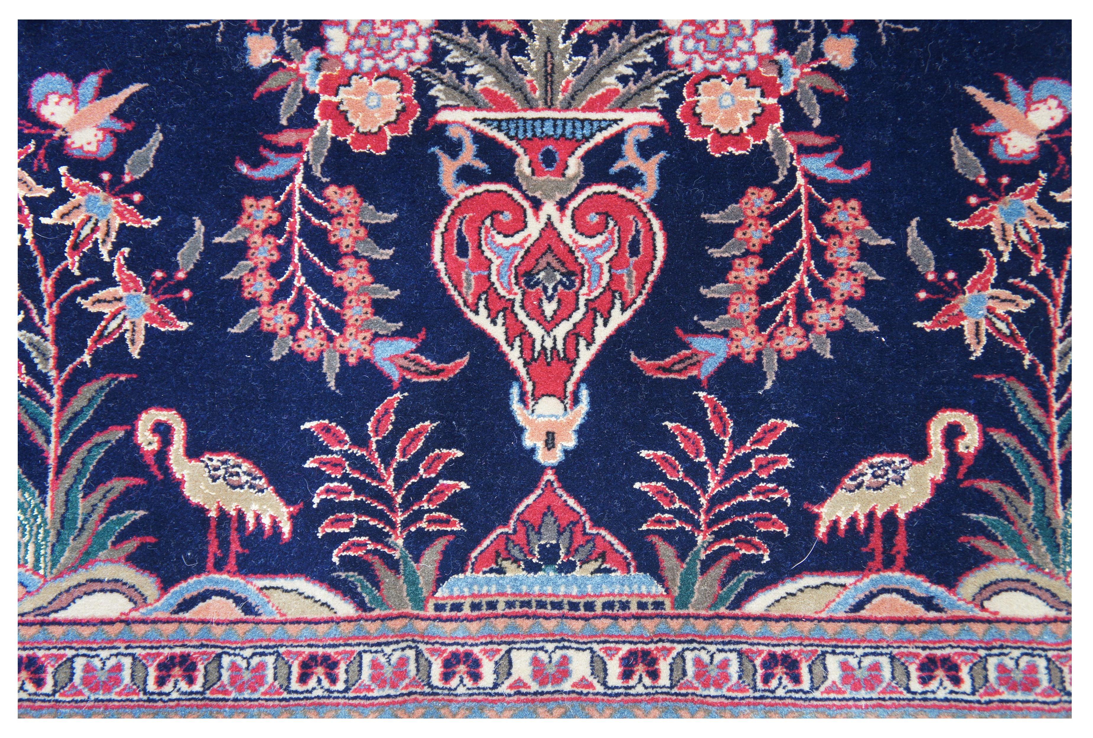 Tapis à vase oriental persan vintage présentant un champ de bleu marine avec un grand vase central composé de fleurs rouges, bleues, vertes, orange et roses. La pièce est rehaussée de papillons et d'oiseaux (flamant rose ou cigogne ? / moucheron à