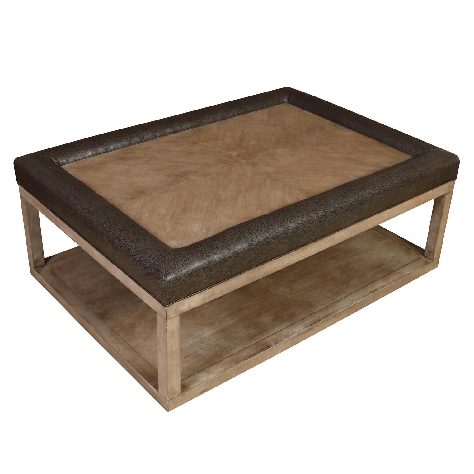 A modern rectangular two tiered coffee table of gray stained wood and brown leather trim detail.