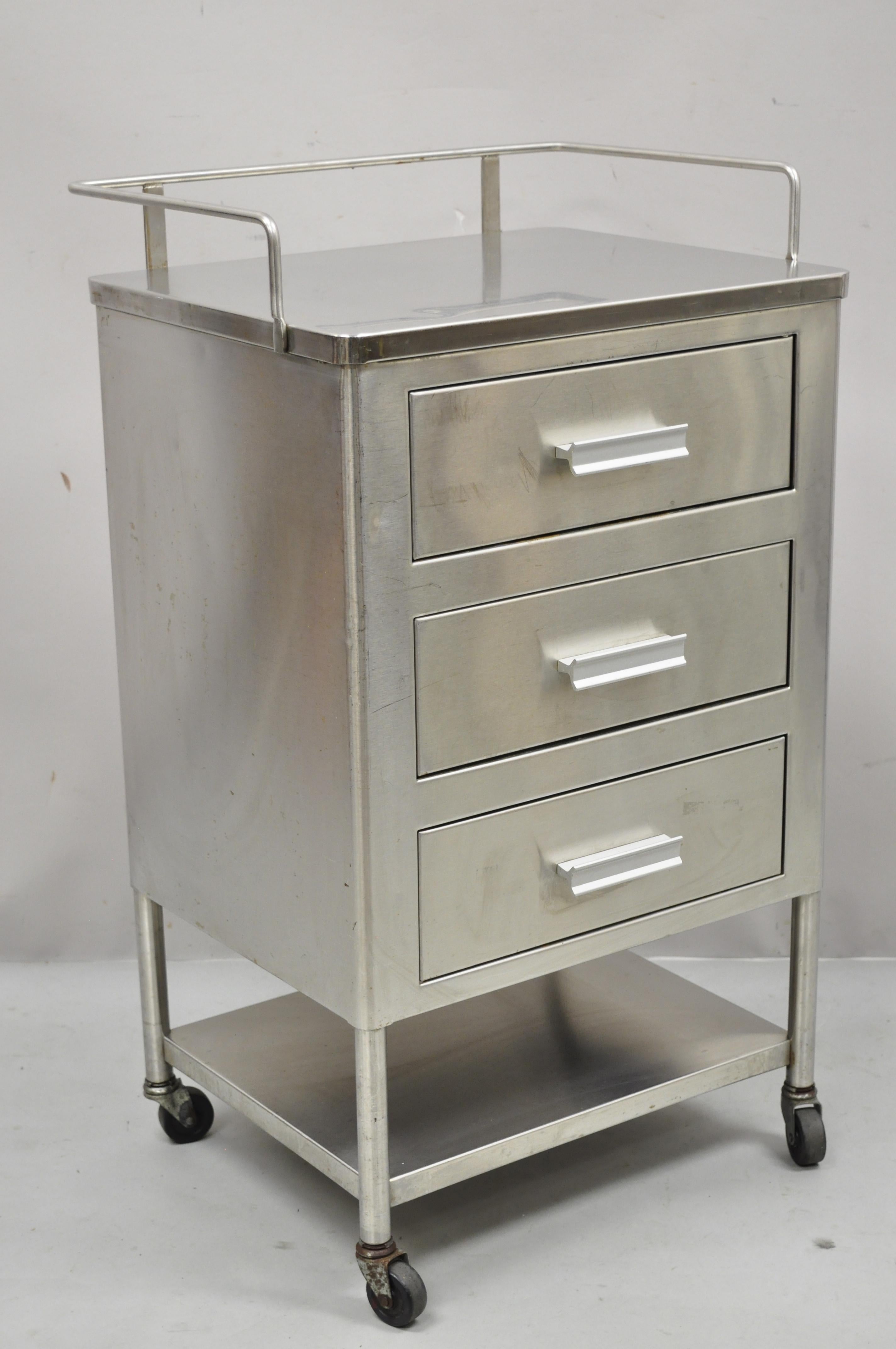 Vintage stainless steel 3 drawer rolling medical dental cabinet nightstand side table on wheels. Item features rolling casters, stainless steel construction, lower shelf, rear gallery, 3 drawers, quality American craftsmanship, great style and form,