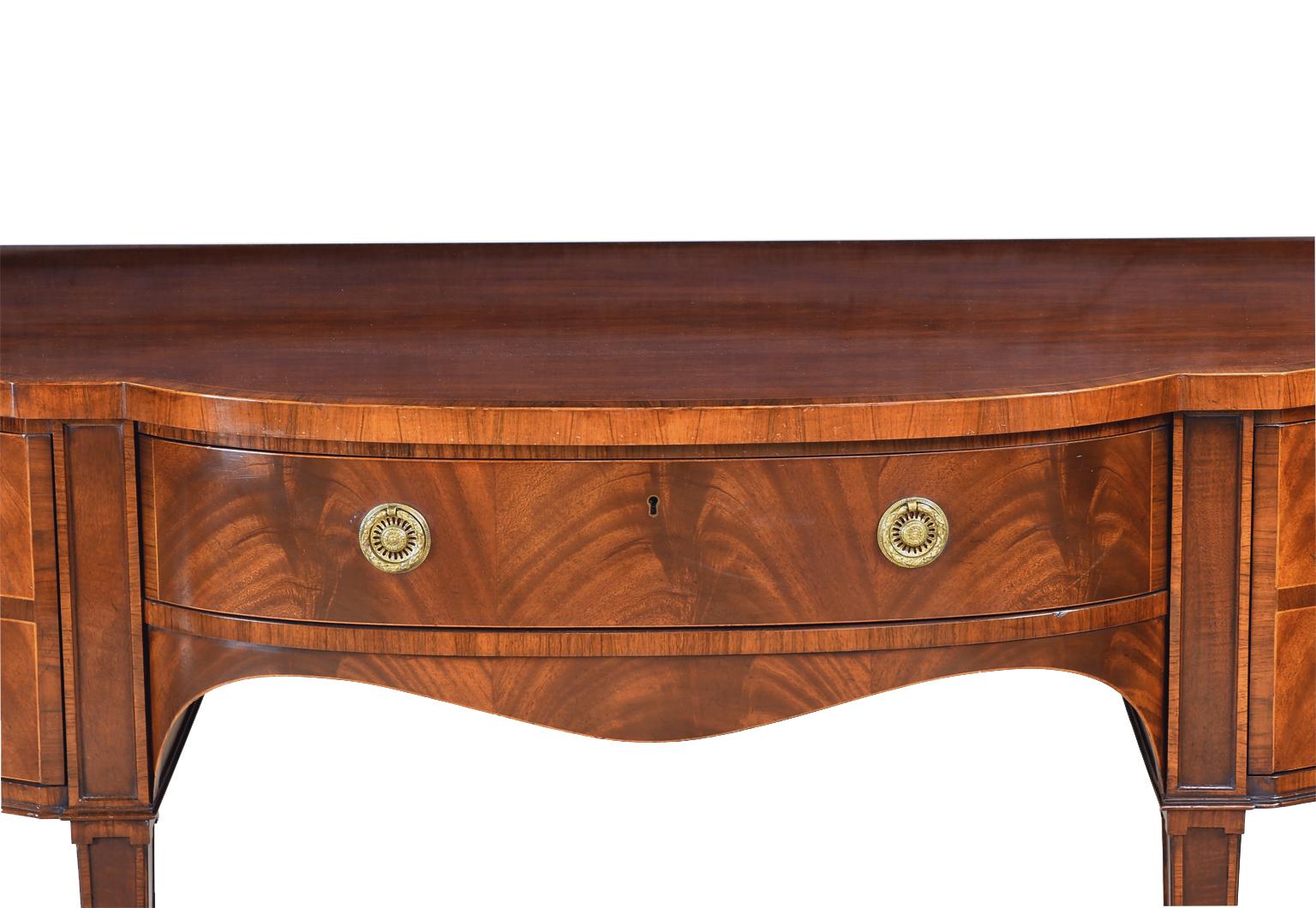 A handsome and well-crafted Sheraton-style sideboard in mahogany manufactured by Baker Furniture Company from their Stately Homes of England and Scotland Collection. Features a serpentine front and sides, with a center drawer for silverware flanked