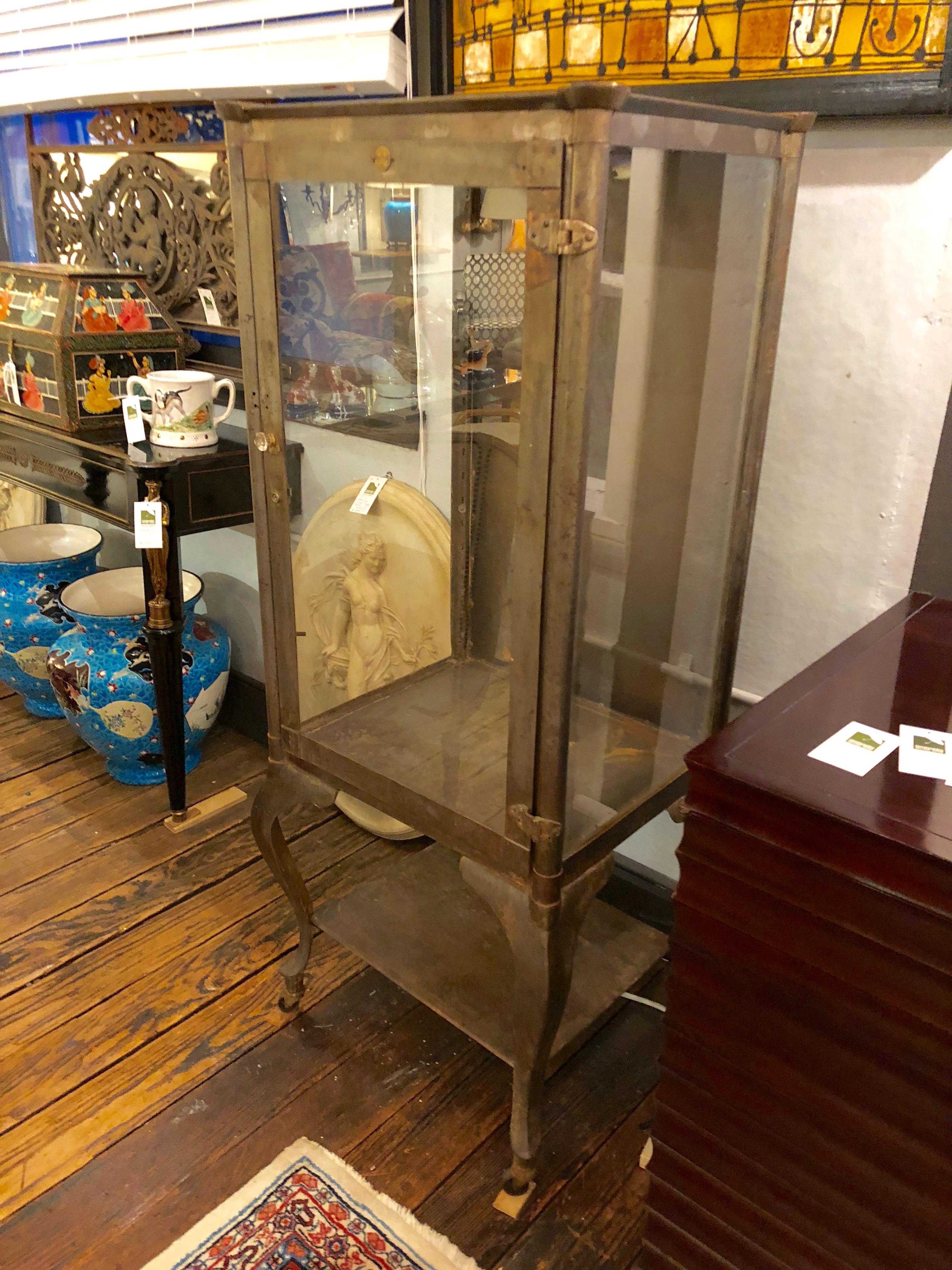 A rare find in a medicine cabinet having stripped grey steel and original glass on front and sides, with elegant curvy cabriole legs on casters. 3 adjustable glass shelves. Brass medallion on the front.