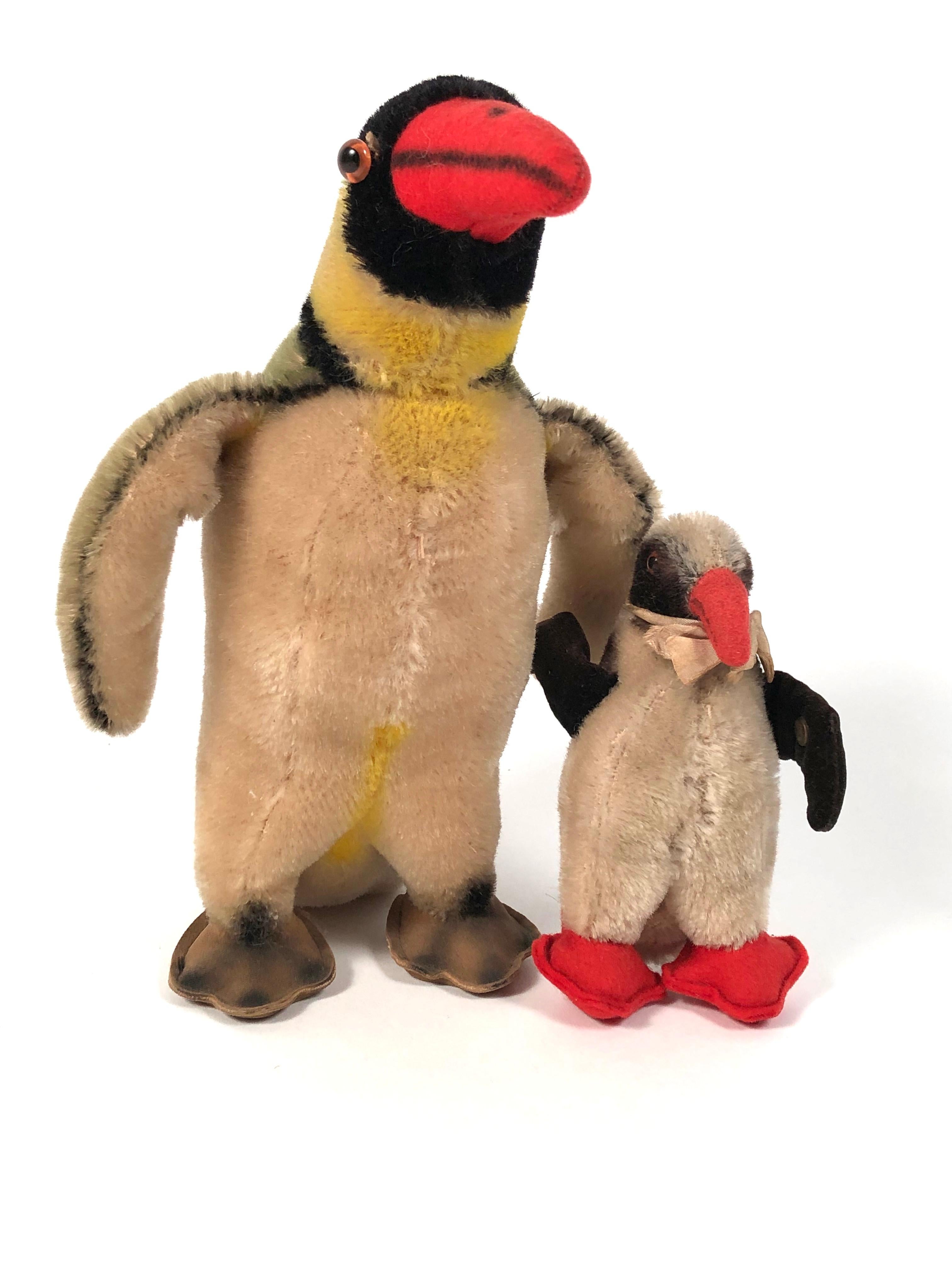 A vintage Steiff penguin and chick, with tan, yellow (penguin) and black bodies and red beaks and feet (chick), both with amber and black glass eyes, the chick with silk scarf tied around its neck. Both with Steiff metal button tags under their left