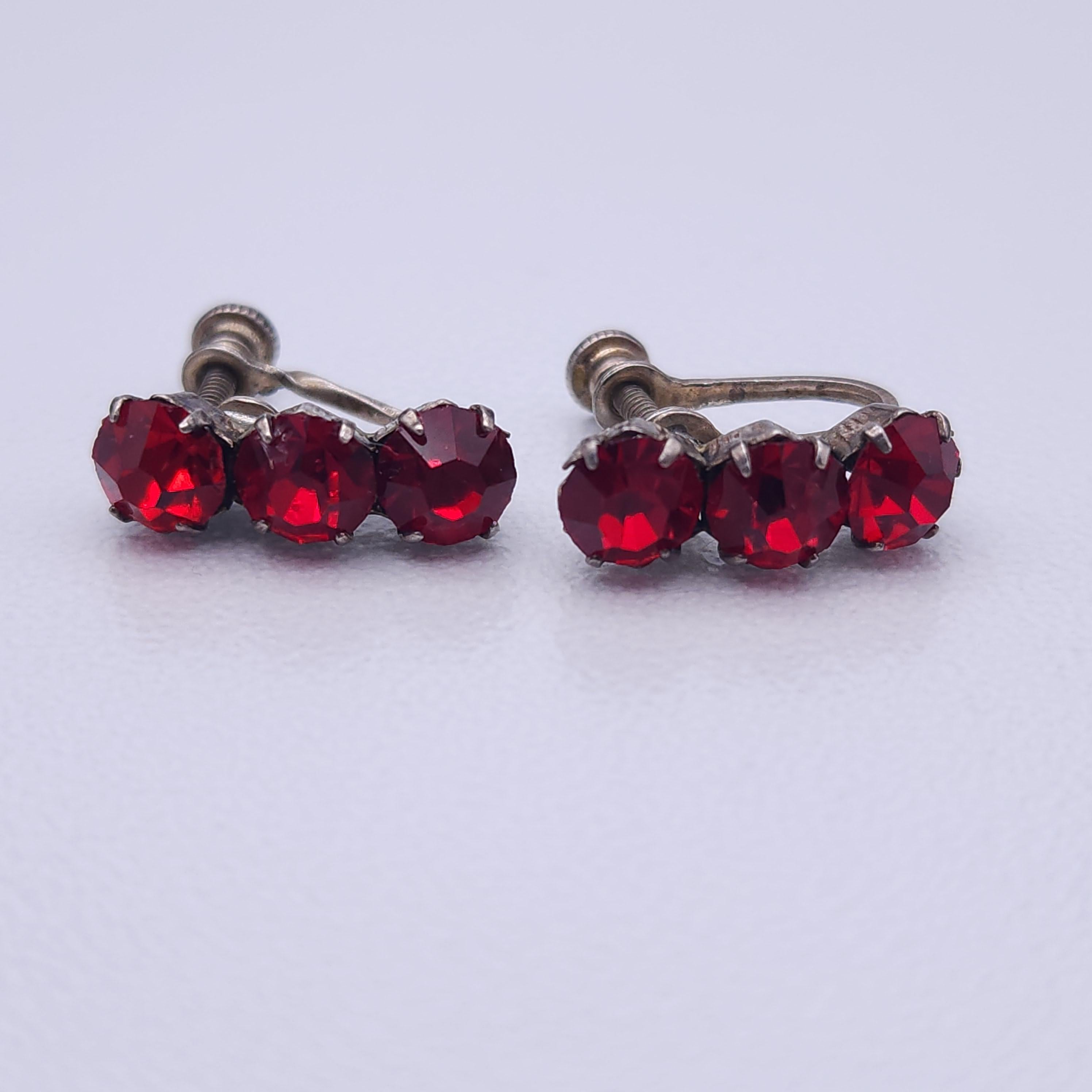 These vintage screw-back earrings feature three vibrant ruby red crystal gemstones in each earring, offering a stunning sparkle that catches the light beautifully. Set in an elegant art deco-inspired silver-tone setting, these earrings exude