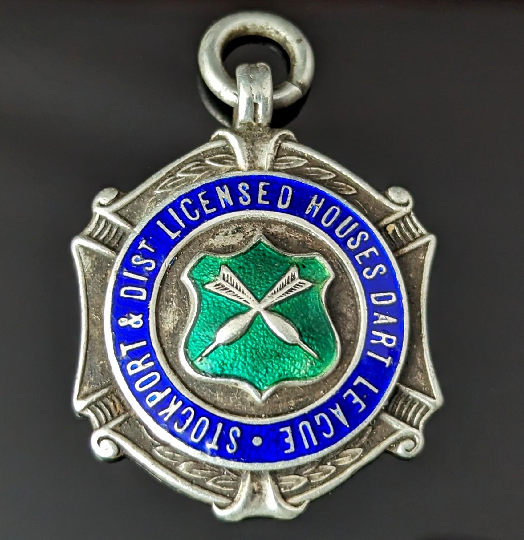 An attractive vintage sterling silver and enamel fob pendant.

It is a nicely shaped piece with an attractive blue and green enamelled design, the green enamel shield shaped centre depicting a pair of crossed Darts.

The text within the blue enamel