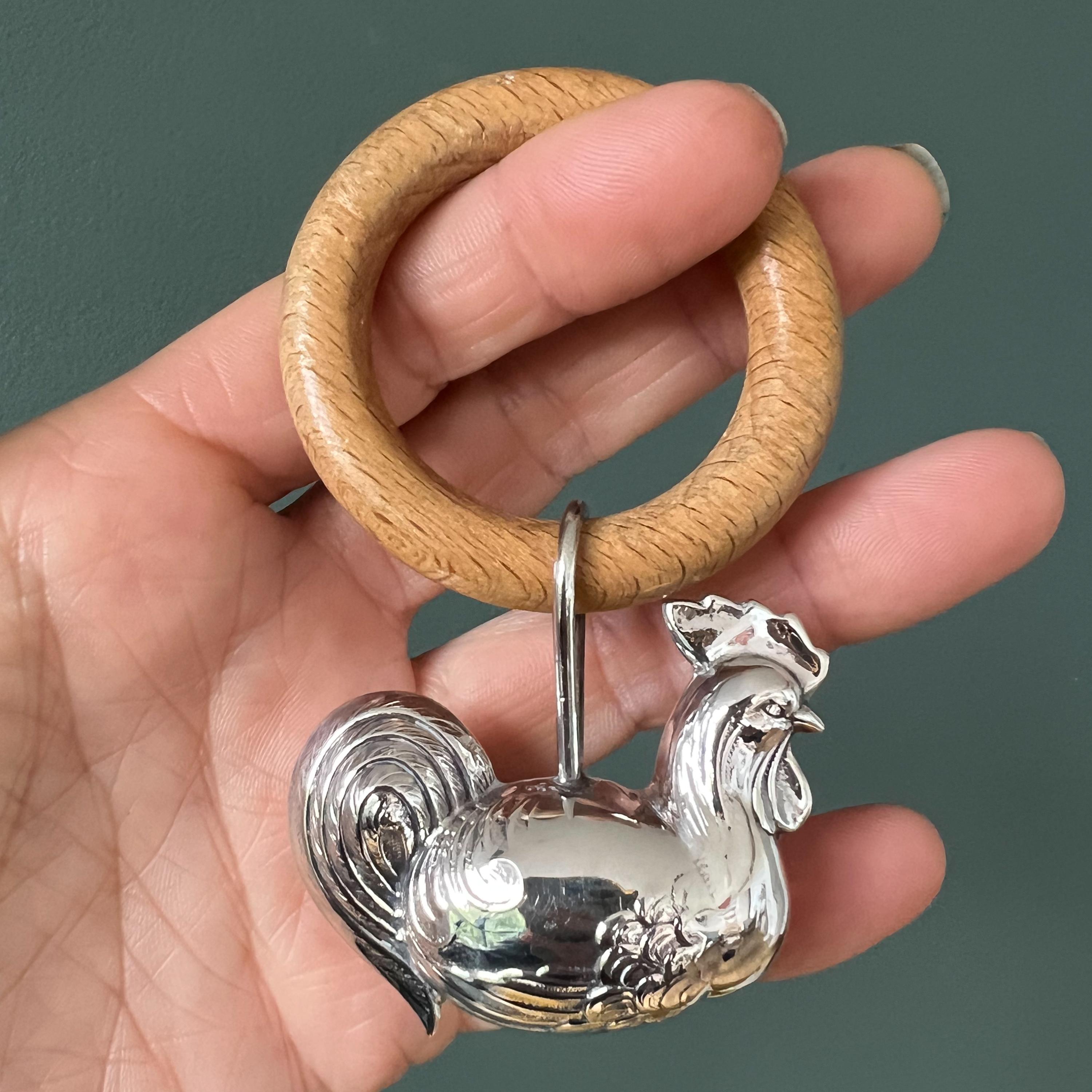 This charming vintage 1950's sterling silver baby rattle is crafted in the shape of a chicken and hangs from a wooden bite ring, making it a meaningful and thoughtful gift for a baby. The chicken is a symbol of nurturing, protection, and new