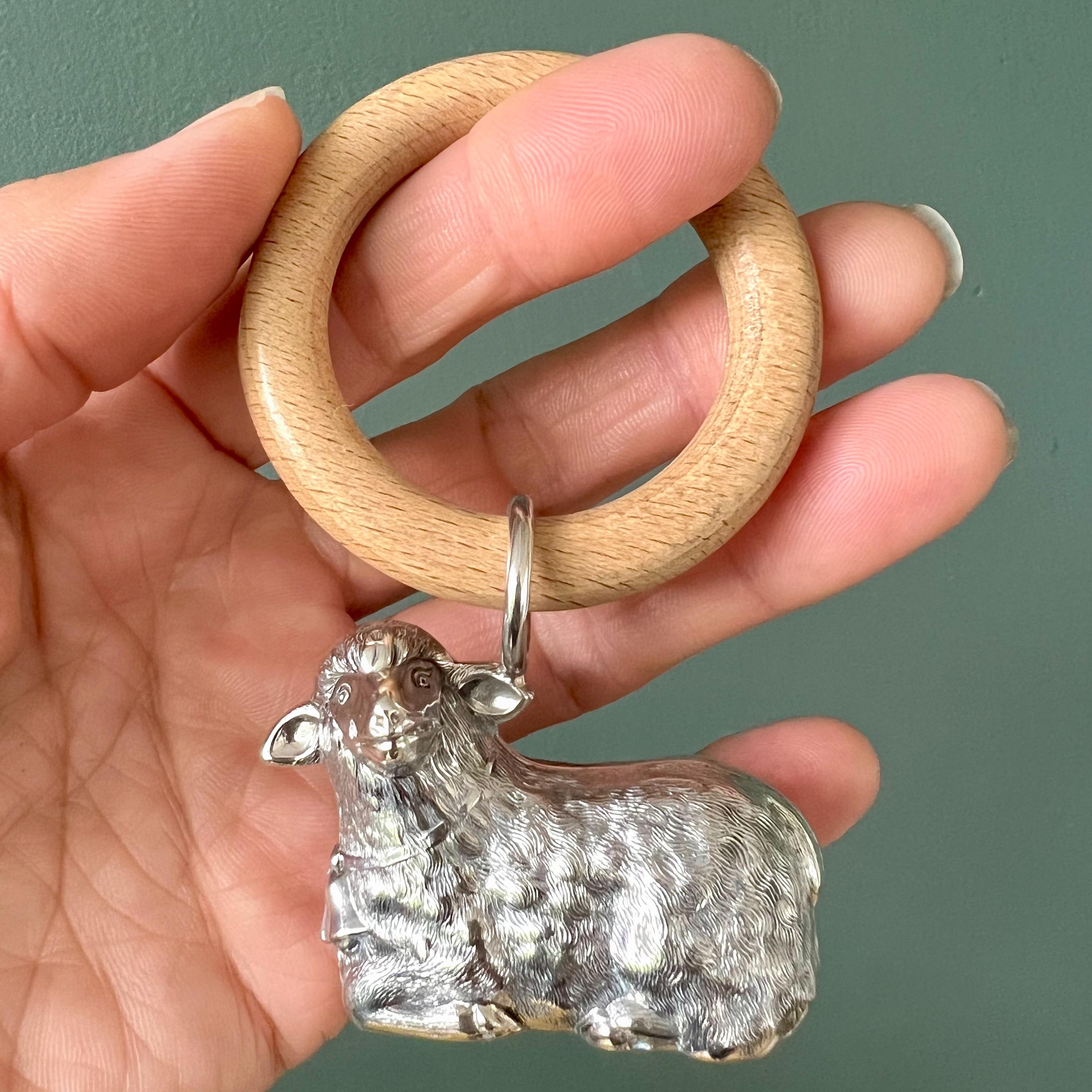 This exquisite vintage 1950's sterling silver baby rattle is shaped like a lying lamb, delicately hanging from a smooth wooden bite ring. The rattle is beautifully detailed, capturing the sweet and gentle features of a lamb, including its adorable