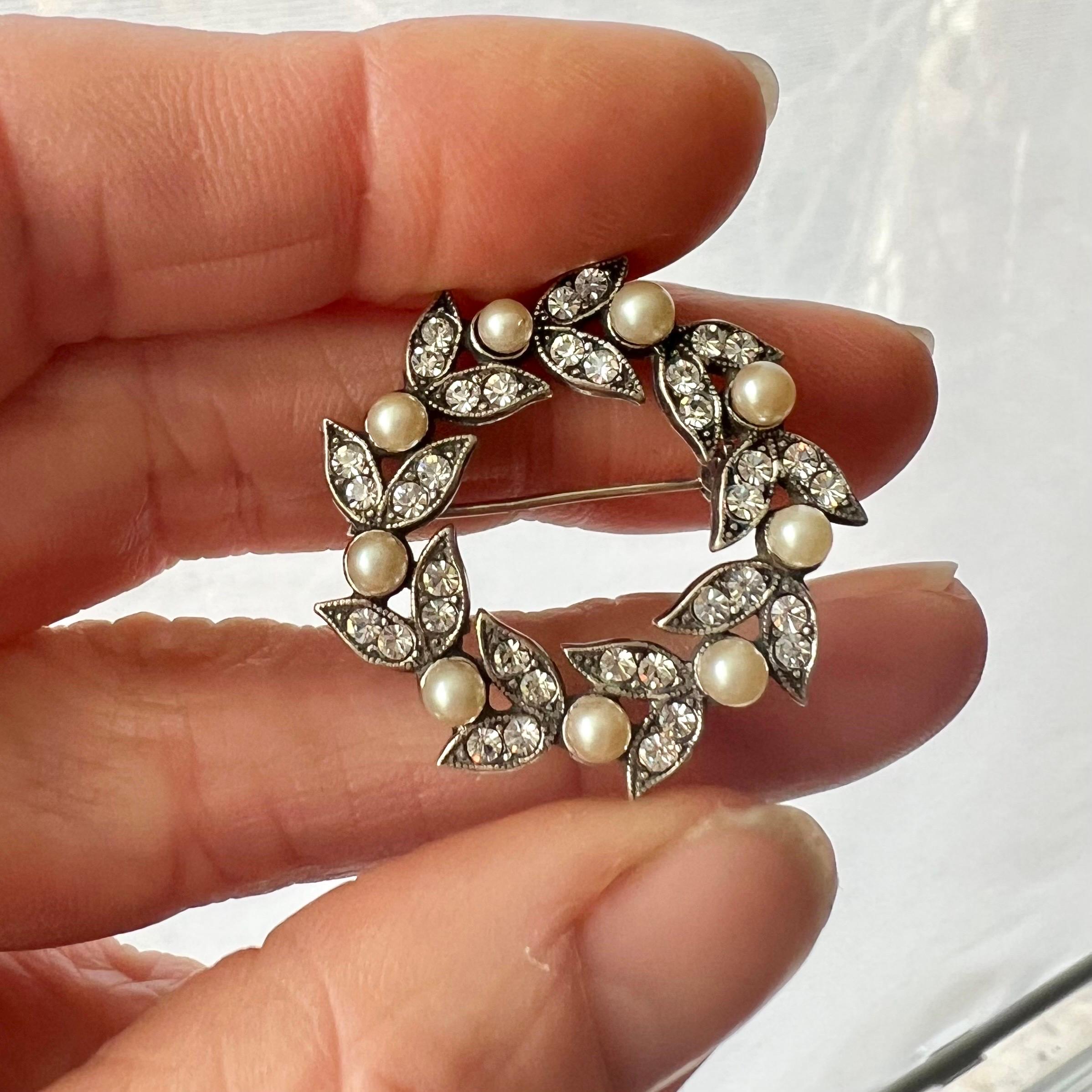 This is a lovely vintage sterling silver round wreath brooch set with faux pearl and glass rhinestones. The brooch is created with diamond-like glass stones also called rhinestones these stones are set in silver shaped petals. The rim of the petals