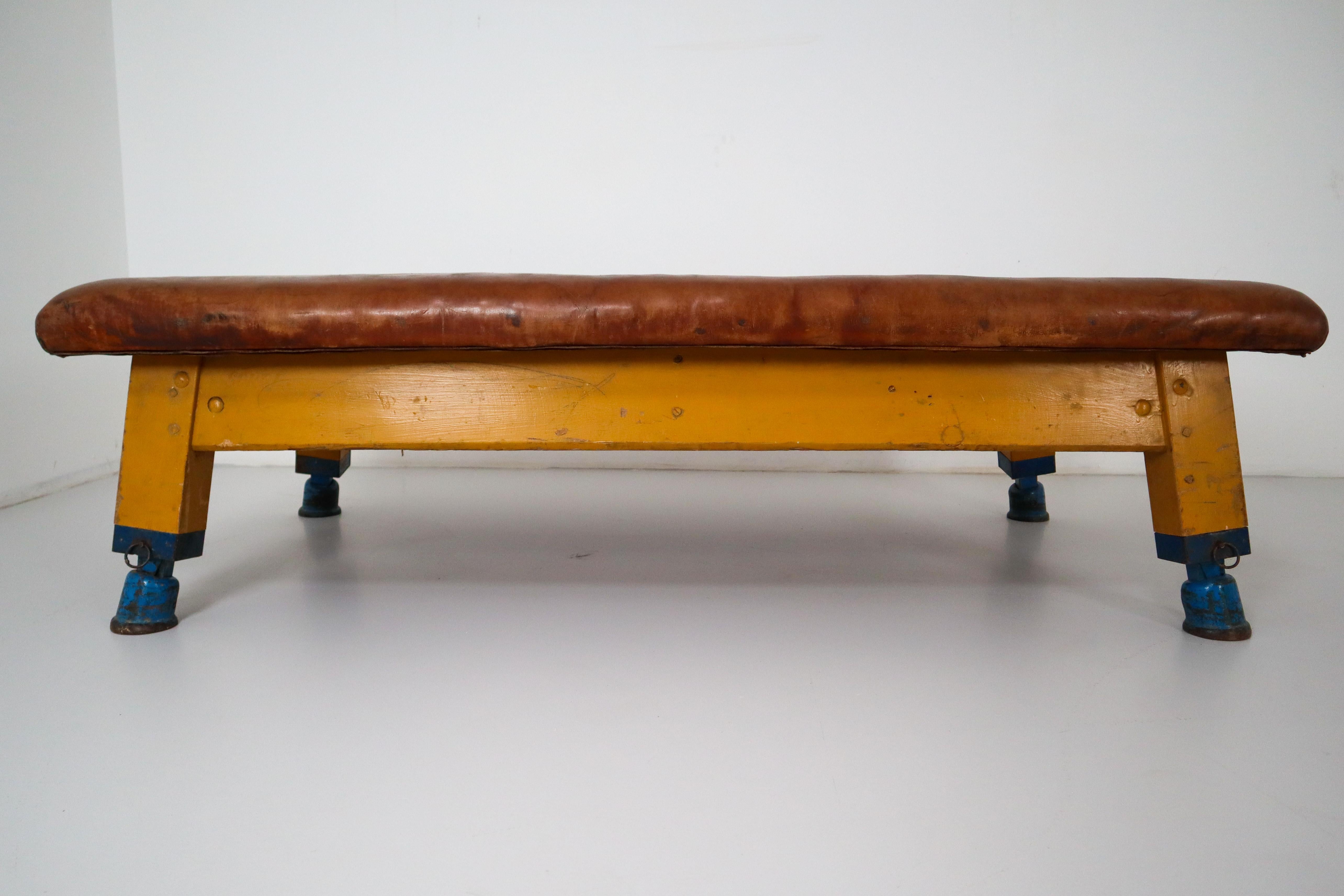 Strong, vintage European gymnasium bench with a patinated leather covered top that sits in to the bench's solid wooden frame. The gym bench frame maintains its original markings where the straps and rings were once held. Each of the four legs has a