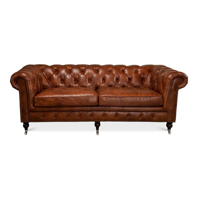 A vintage-style Classic English Chesterfield leather upholstered sofa, with top-grain leather in a vintage brown finish. With tufted backrest and rollover arms, with two padded cushions raised on turned mahogany feet with brass casters and finished