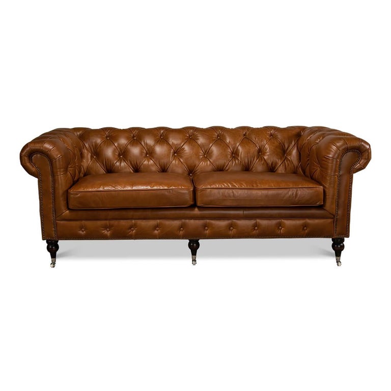 A vintage-style classic English Chesterfield leather upholstered sofa, with top-grain leather in a vintage Cuban cigar color. With tufted backrest and rollover arms, with two padded cushions raised on turned mahogany feet with brass casters and