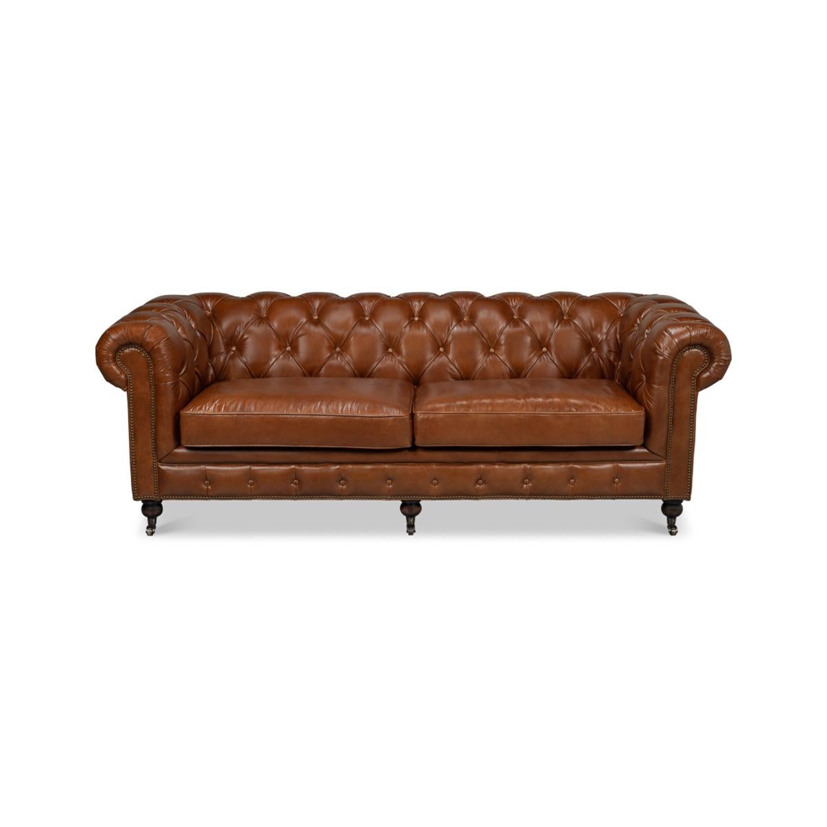 A vintage style Classic English Chesterfield leather upholstered sofa, with top-grain leather in a Havana brown finish. With tufted backrest and rollover arms, with two padded cushions raised on turned mahogany feet with brass casters and finished