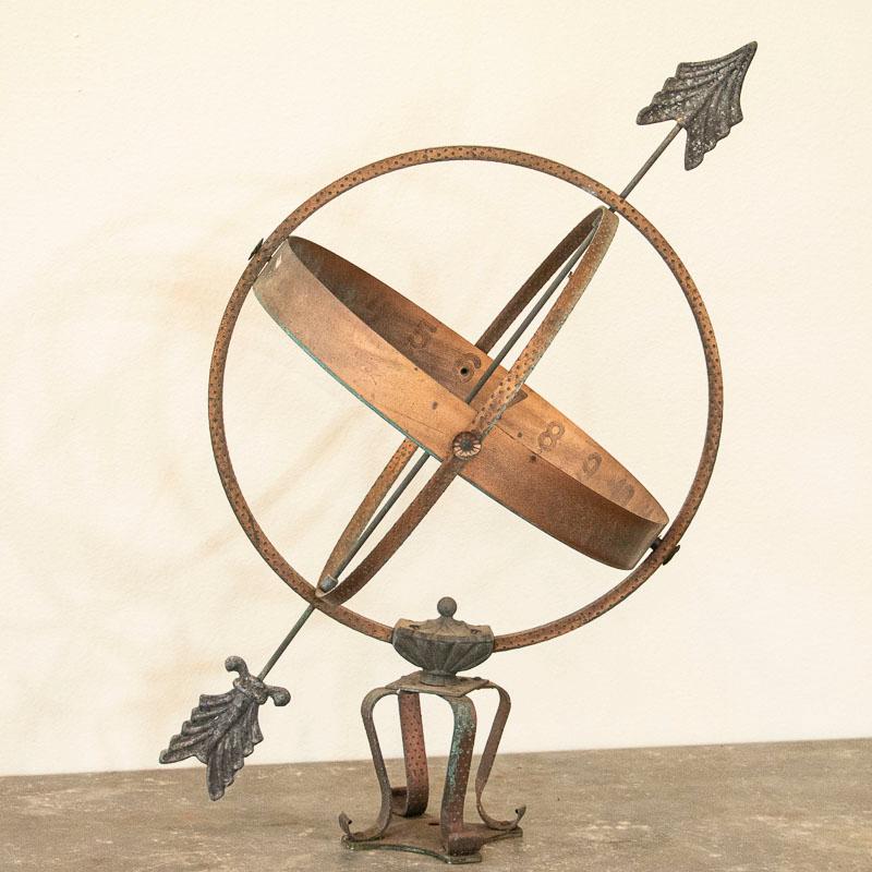 This vintage copper and vertigris globe shaped garden ornament, called an armillary, is made up of metal bands, pierced by an arrow and supported by a mounting bracket. The base resembles a crown while the fitting is a bit loose where the globe