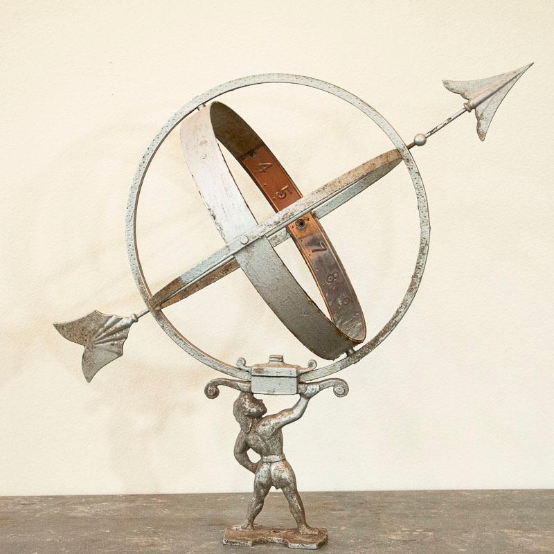 This vintage silver painted globe shaped garden ornament, called an armillary, is made up of metal bands, pierced by an arrow and supported by a mounting bracket. This has a favored traditional figure of Altas holding up the world or globe. These