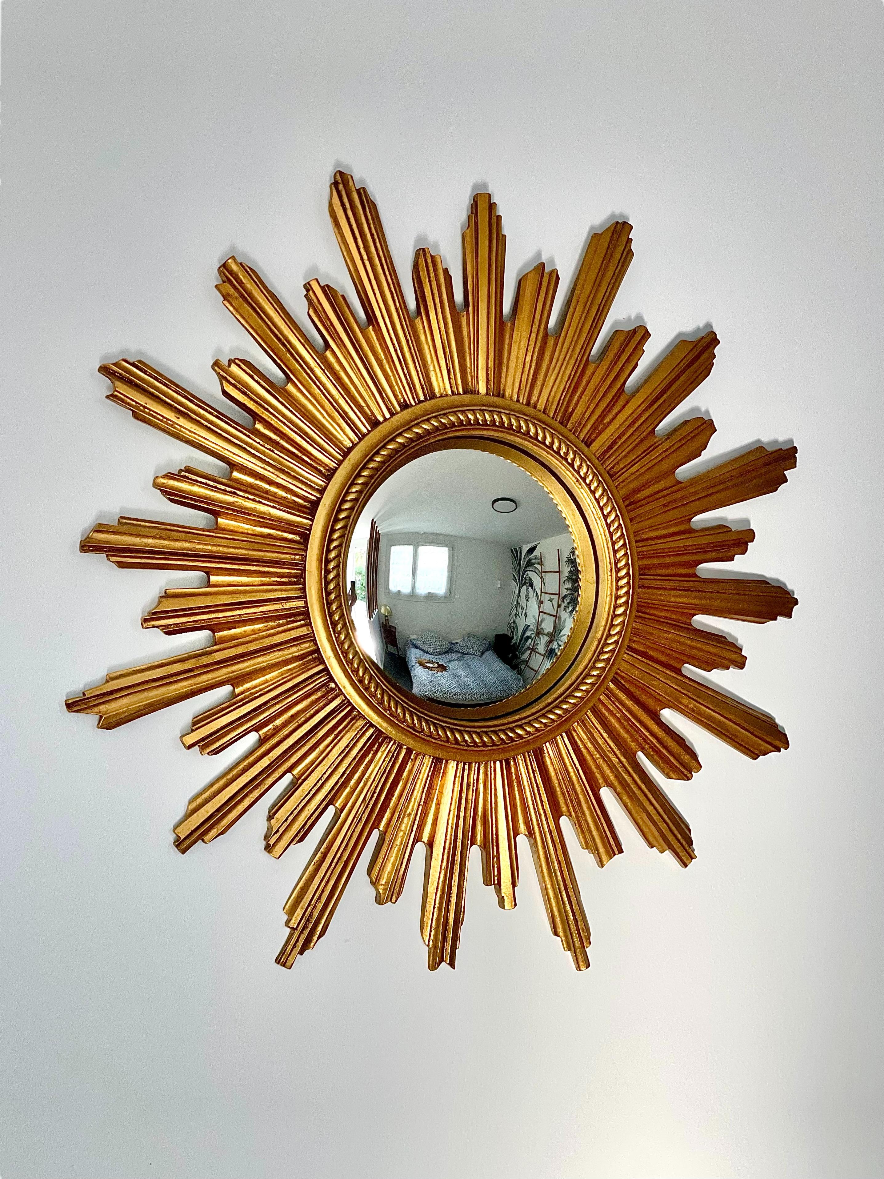 Ce superbe miroir mural vintage à effet soleil présente une plaque de miroir convexe, entourée d'un ensemble de tiges en bois sculpté et doré, qui imitent les rayons du soleil. La plaque de miroir elle-même est bordée d'un cadre classique de perles