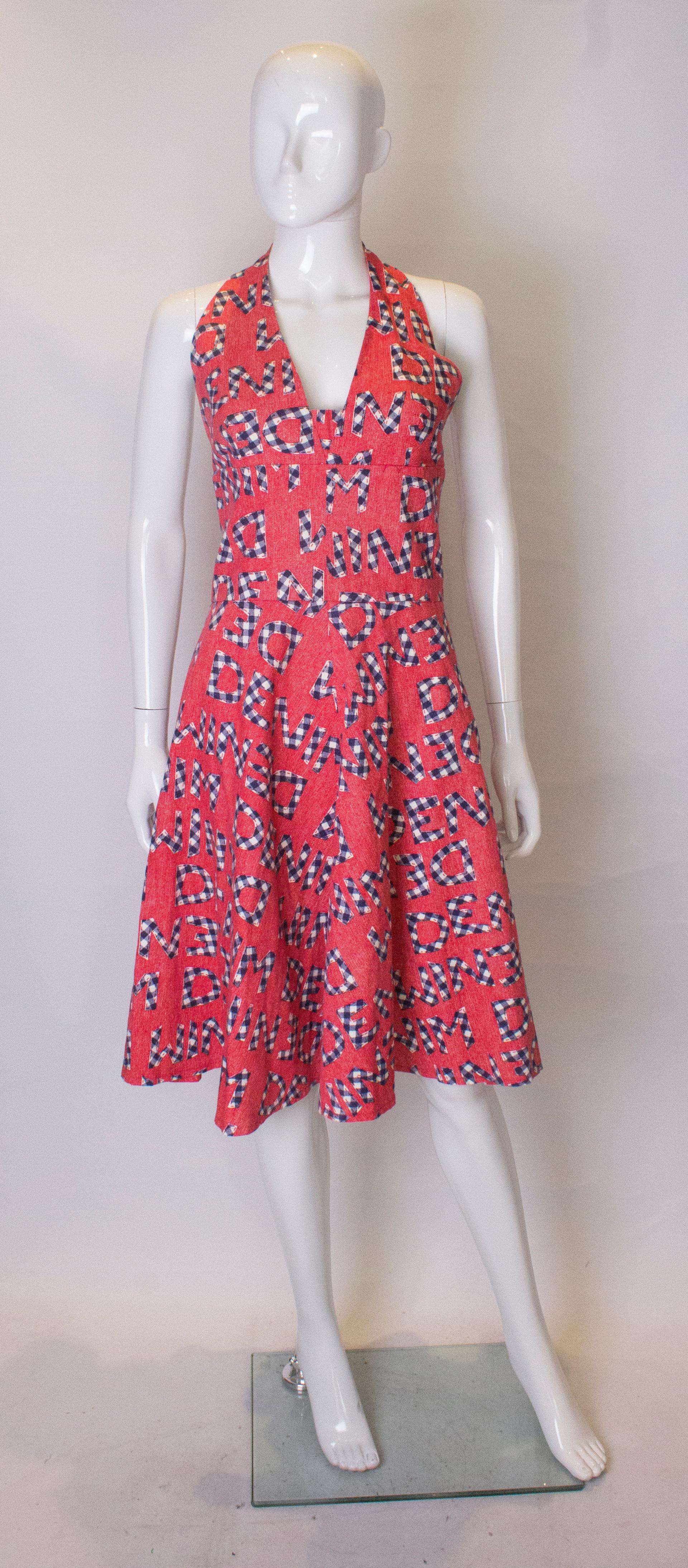 A fun summer dress by Susan Small.  The dress has a halterneck and flared skirt with a zip opening. The fabric has a red background , and the word 'denim' printed on to it. 