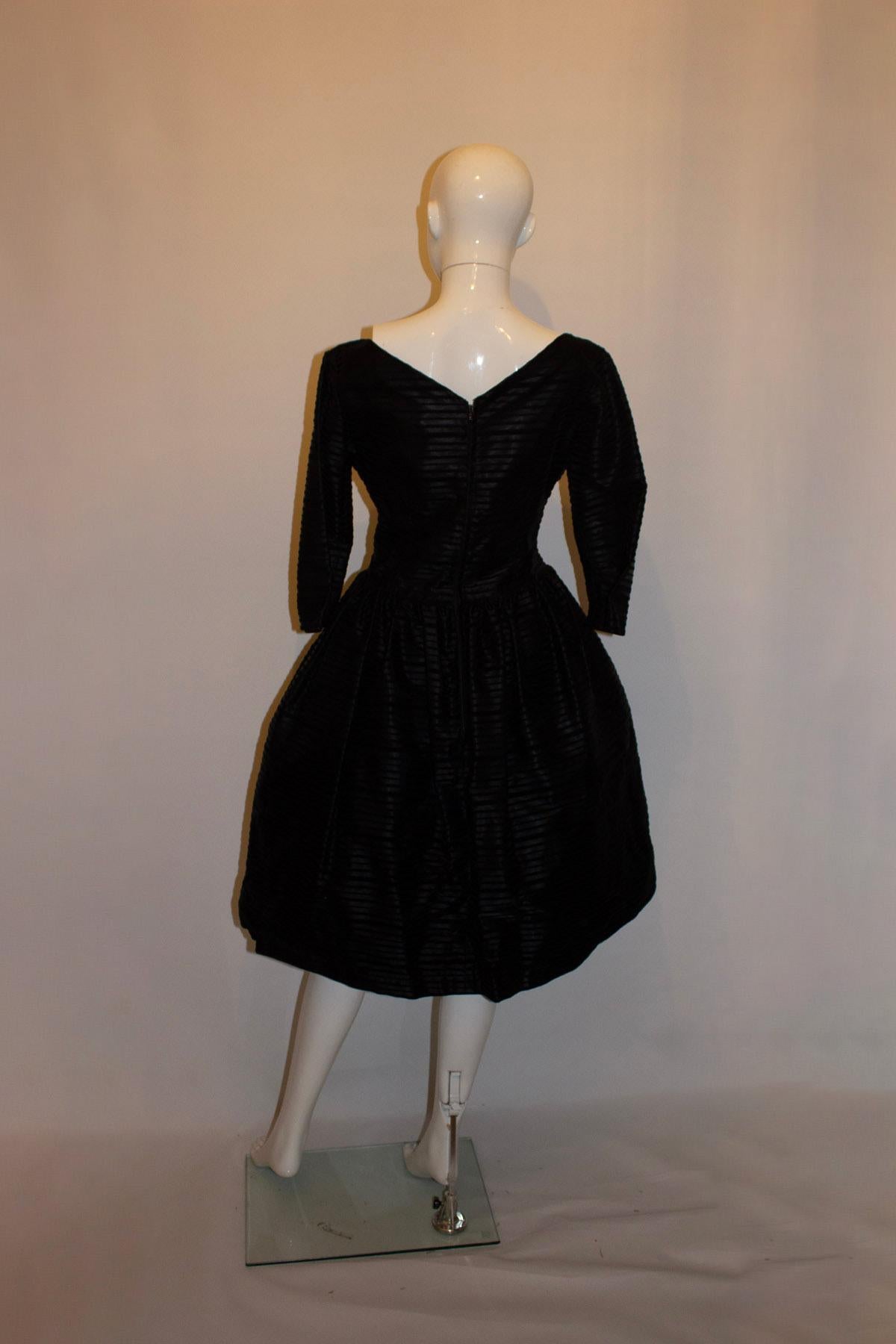 Un elegante vestido de cóctel vintage de Susan Small.  El vestido es negro con bandas horizontales de terciopelo negro. Tiene enagua incorporada, abertura trasera con cremallera, escote barco bajo delante y una profunda V detrás. El vestido tiene