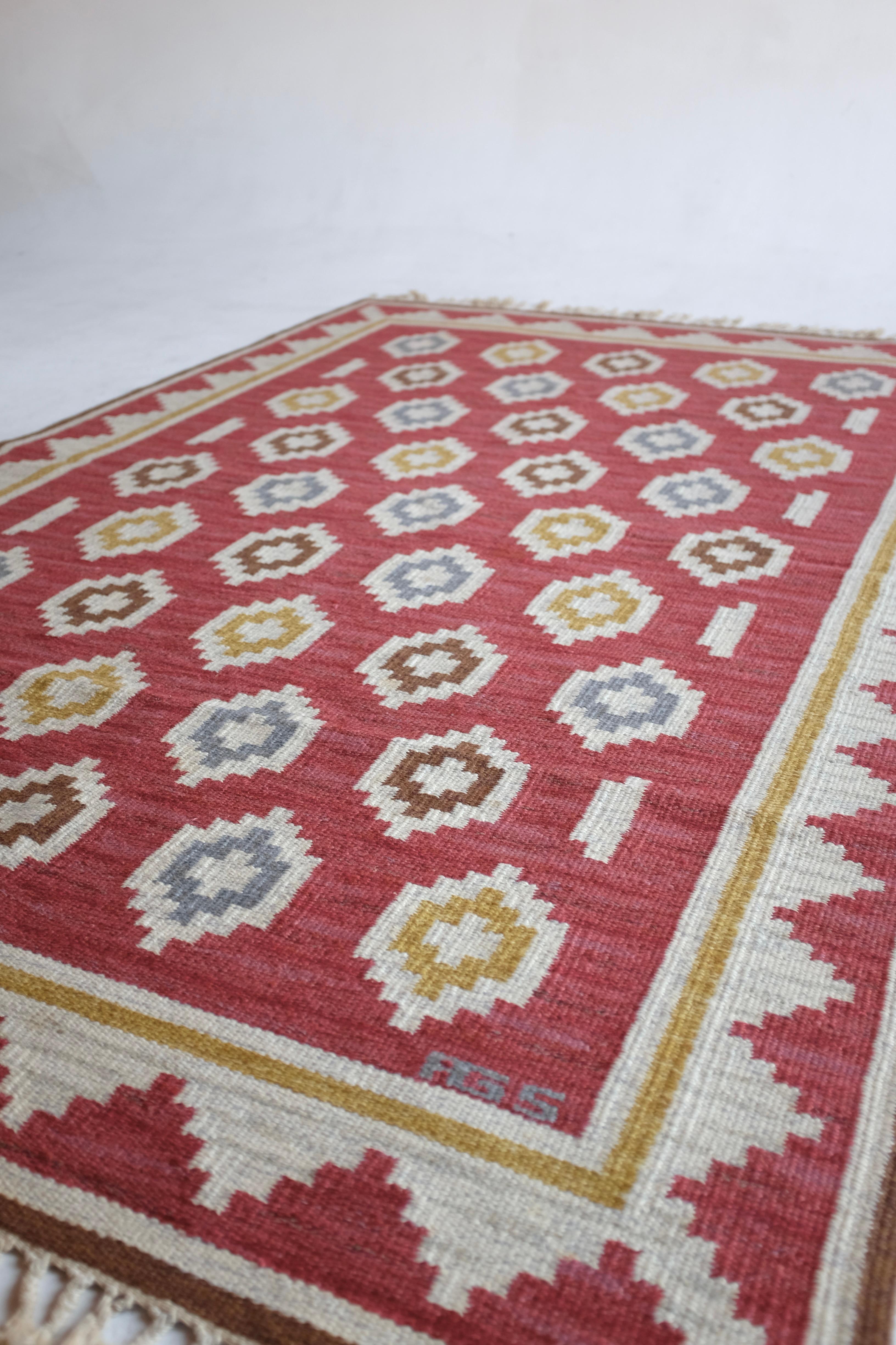 Magnifique tapis suédois vintage Rug & Kilim de la designer Anna Greta Sjöqvist. Rouge vif sur fond blanc et marron, avec un motif géométrique de fleurs en jaune, bleu et marron. Initiales du designer en bas à droite. Bon état vintage avec toutes