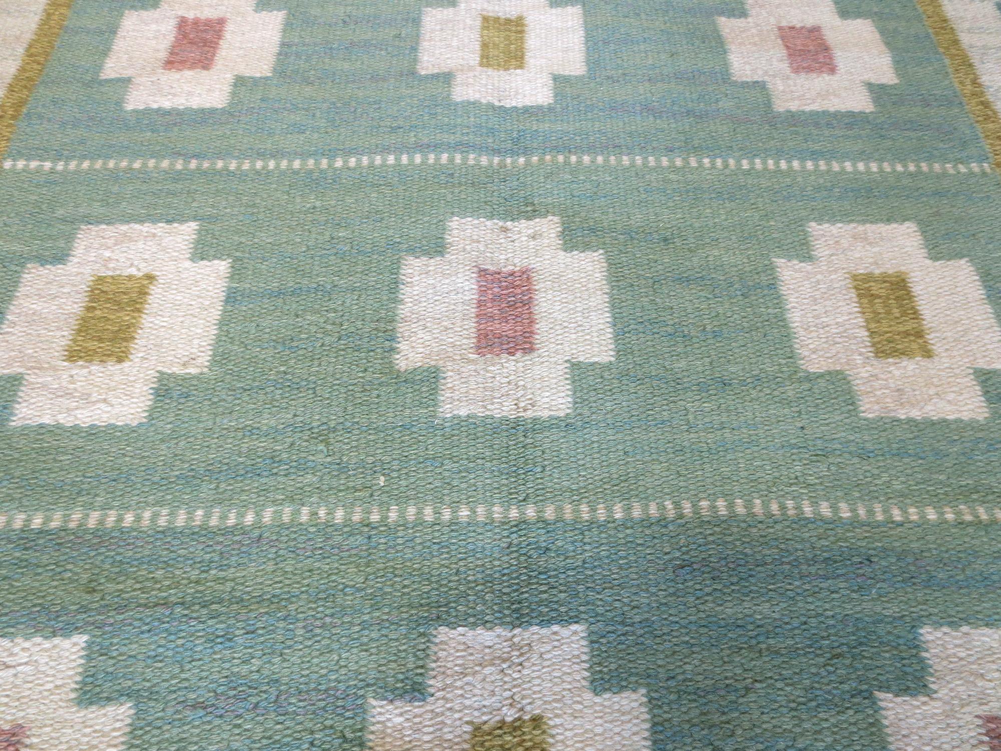 This is a vintage Swedish Kilim signed by artist Anna Greta Sjoqvist from the mid-20th century. It features calming tones of greens, greys, ivories, rosy pinks, and yellows in repeating geometric motifs that runs within the borders and set against