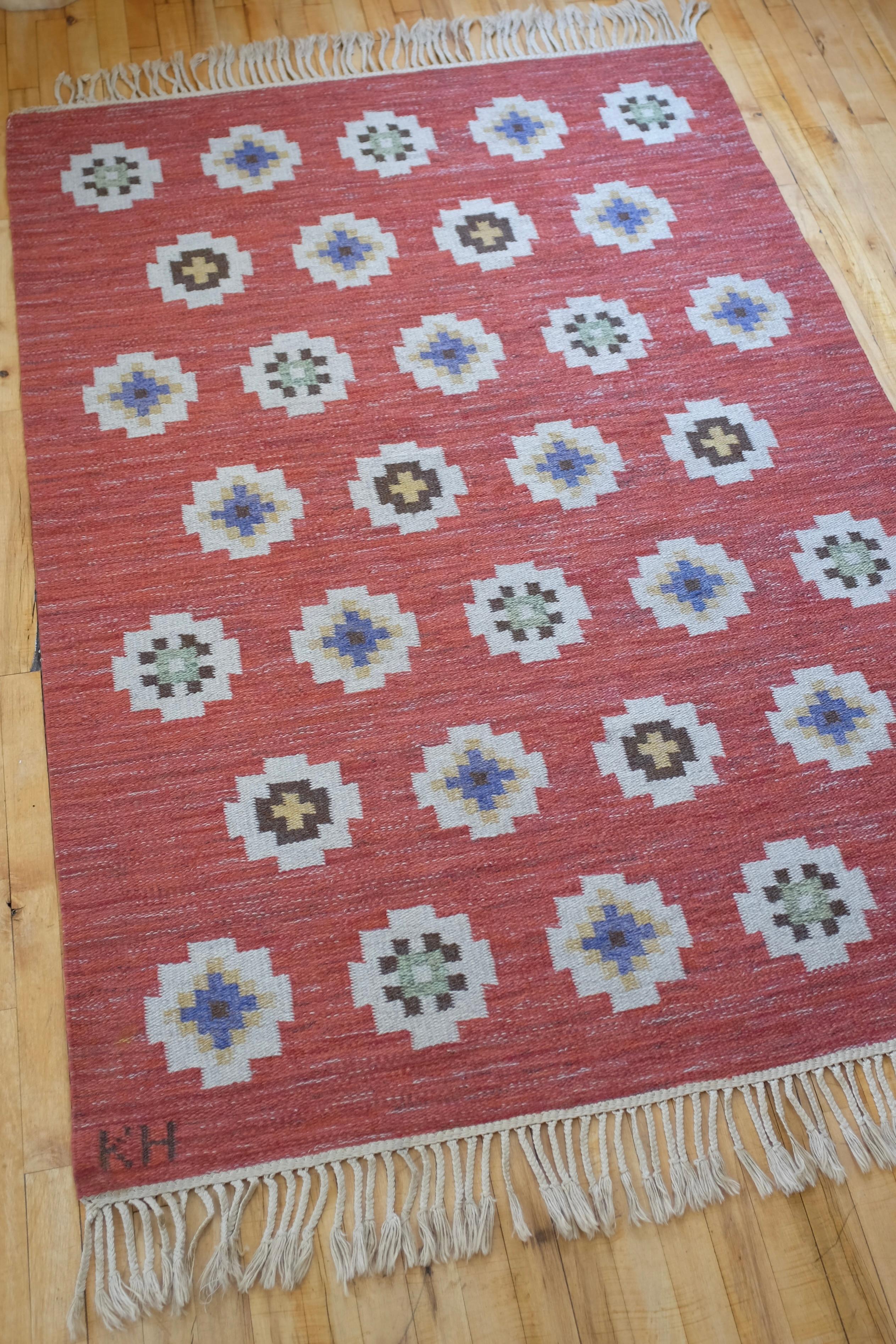 Splendido tappeto vintage svedese di metà secolo a motivi geometrici con le iniziali del designer KH. Sfondo rosso con disegno bianco raffigurante fiori con accenti di colore beige, blu, verde e marrone. Condizioni molto buone con la maggior parte