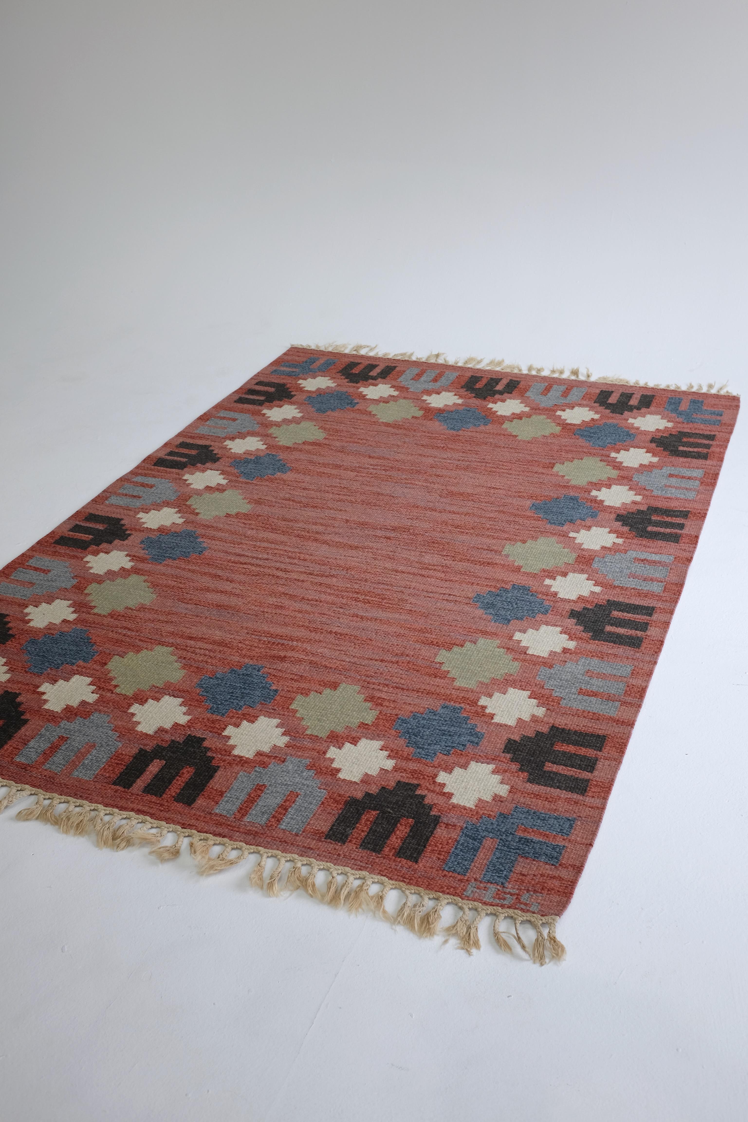 Beautiful Vintage Swedish Kilim rug by designer Anna Greta Sjöqvist. Vibrant pink and red background with a border design of black, blue, white and green geometric flowers. Designer initials on the lower right hand side. Fringes have been worn down