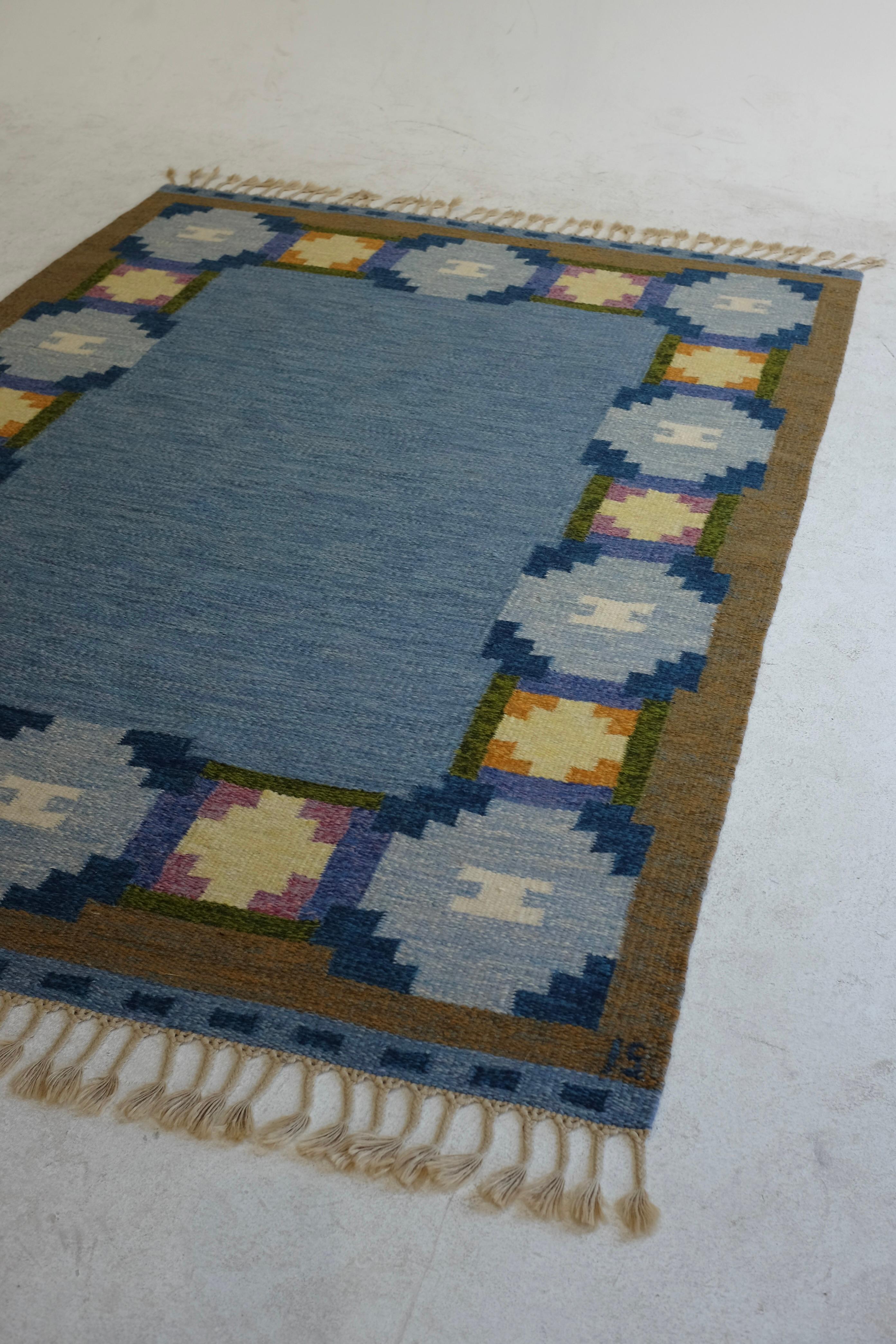Vintage Mid-Century rug by Swedish textile designer Ingegerd Silow. A blue base in the centre is surrounded by a geometric flower design in blue, green and pink with a brown border. Designer initials at the lower right hand side. In a very good