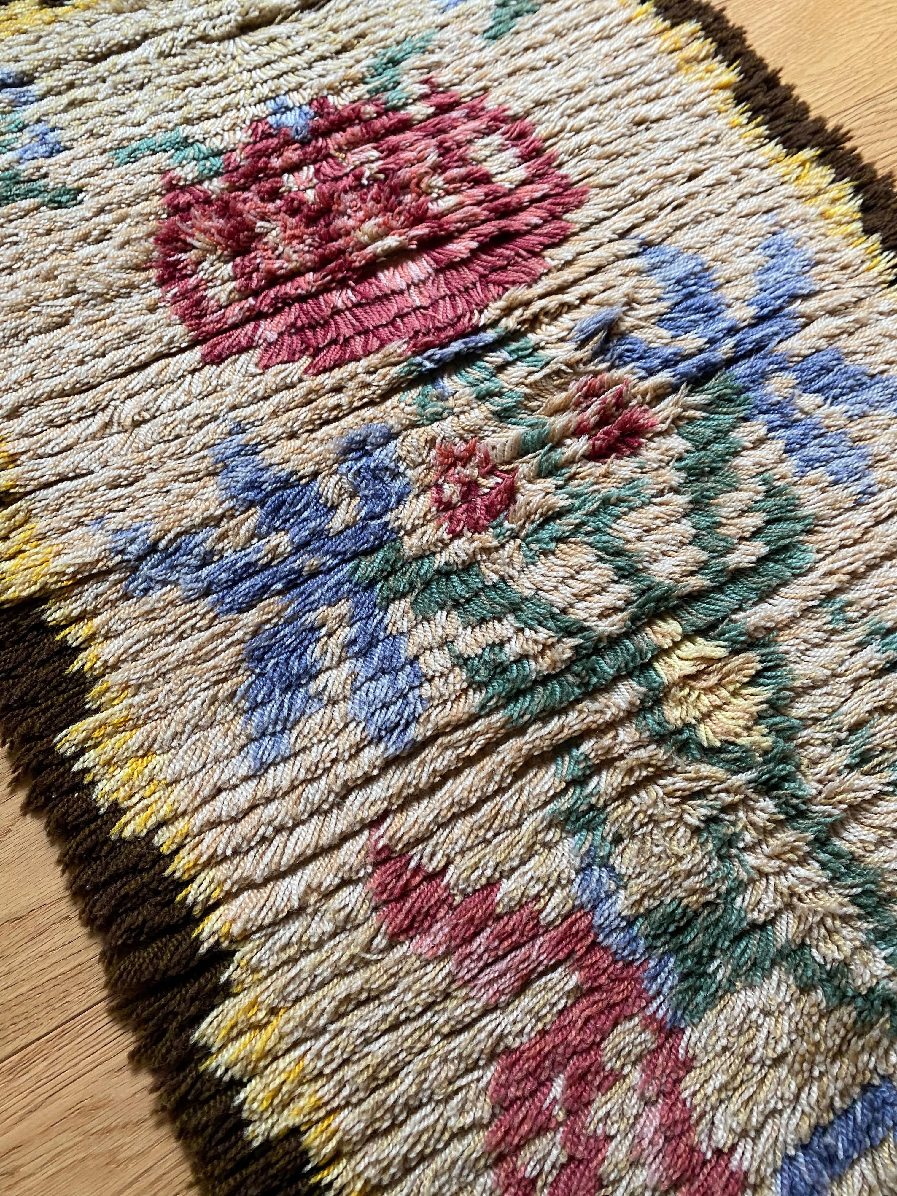 Beautiful small vintage Rya rug from 1938. Compromising of a central flower motif in pink, blue and green with a beige background and brown border. Rya rug or Knotted Pile carpet were popular in Scandinavia as decorative objects to use as rugs, bed