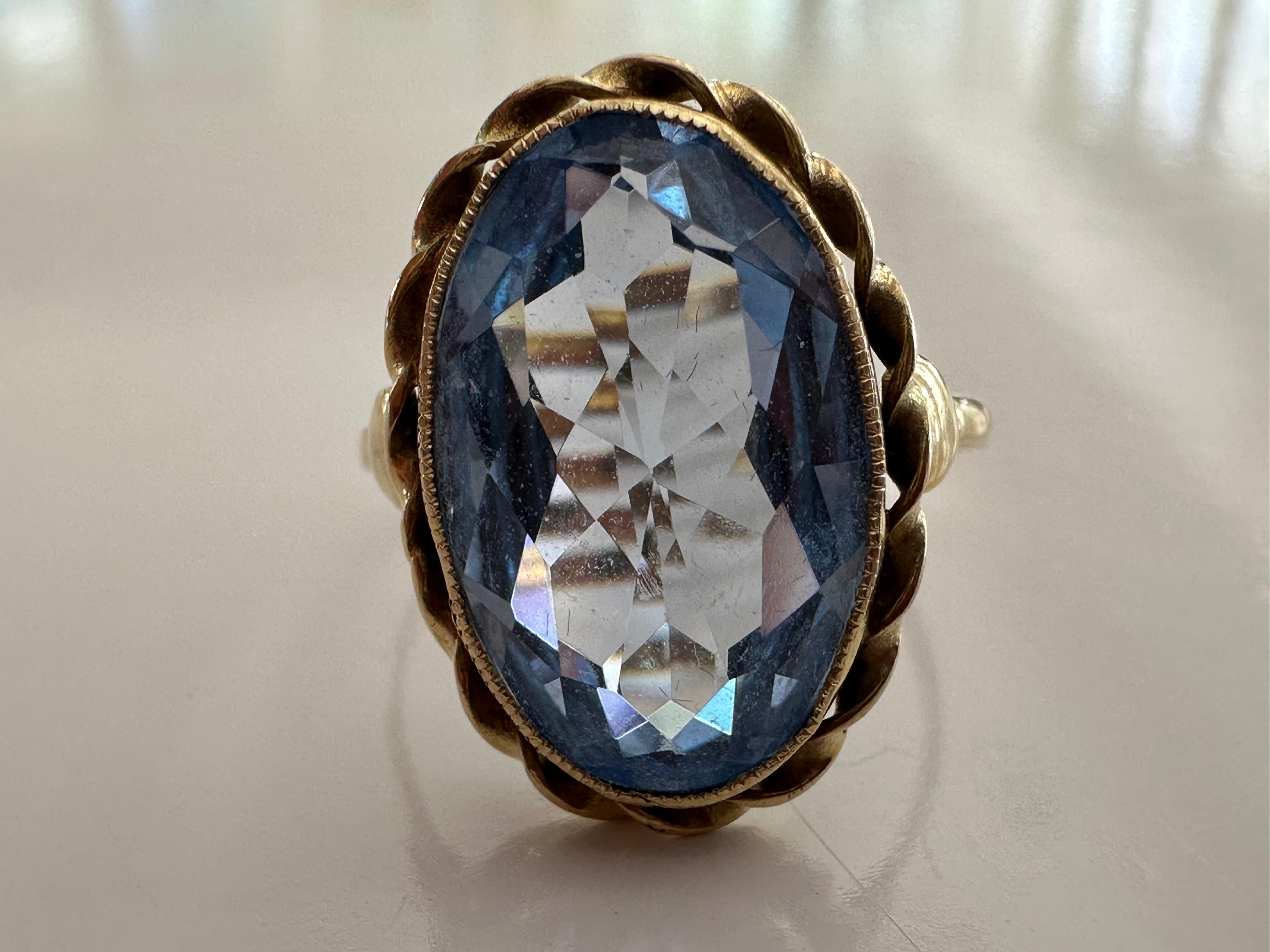 A stunning oval shaped synthetic light blue spinel radiates from within a delicate band fashioned from 14kt yellow gold encircled by a rope design and milgraining. 
