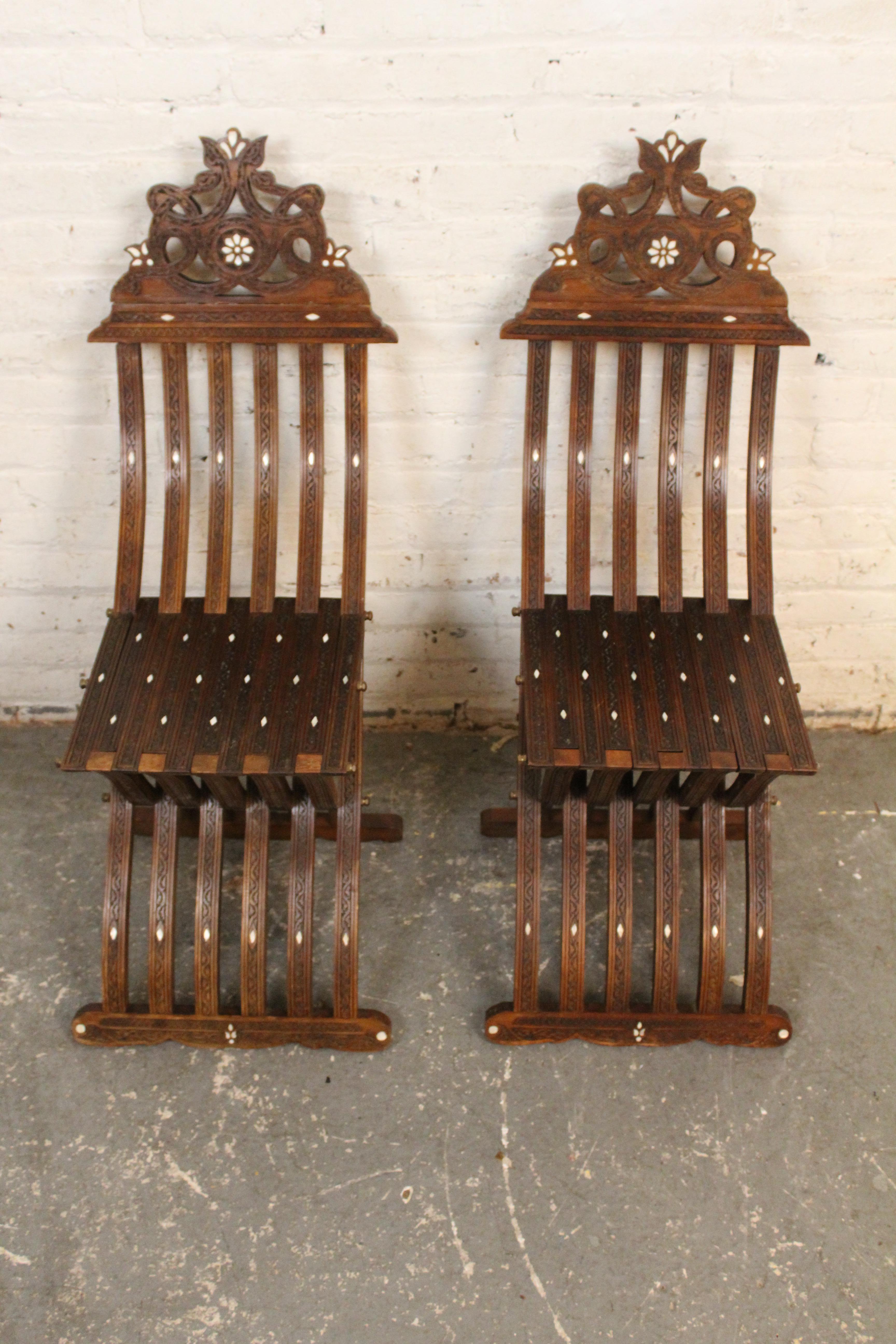 Infondi in qualsiasi stanza un'ipnotica eleganza d'altri tempi con questa squisita coppia di sedie vintage moresche pieghevoli. Realizzate in legno massiccio intricatamente intagliato e con lussuosi intarsi in madreperla, queste sedie pieghevoli a