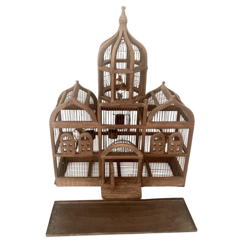 This vintage architectural bird cage has a pair of peaked roofs and tall center dome under which is above a portico leading to a swinging door. The frame is stained wood and is interspersed with metal wire caging. To the left at the bottom is a