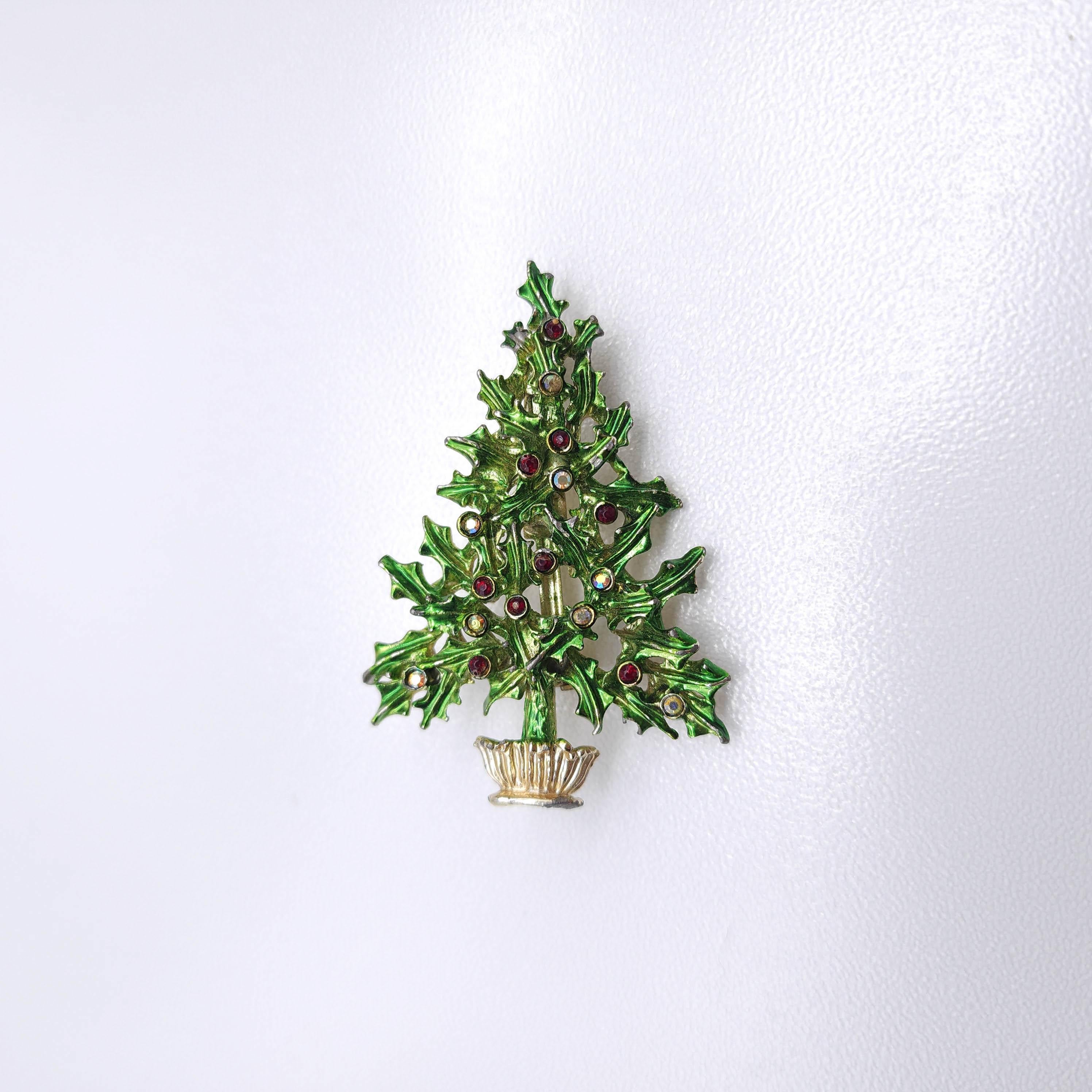 This vintage Christmas tree brooch by Tancer II features a detailed holly leaf design, crafted in green enamel with gold-tone accents. 

The tree is adorned with red and clear  aurora borealis crystals, resembling festive ornaments. 

The textured