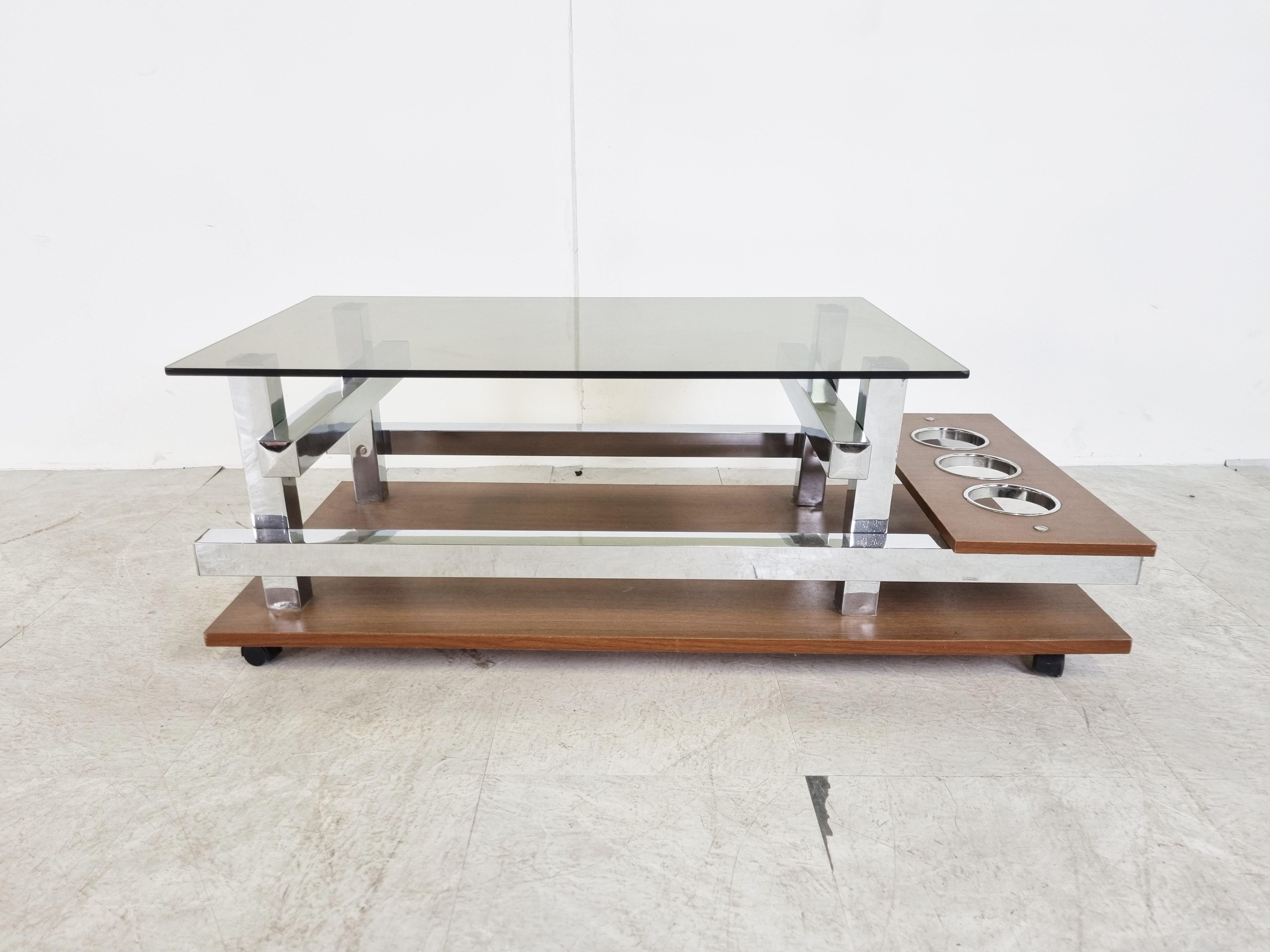 Mid century coffee table made from teak and chrome base with a smoked glass top.

It has an attractive geometrical design and a bottle holder.

1960s - Belgium

Good overall condition

Dimensions:
Height: 36cm/14.17