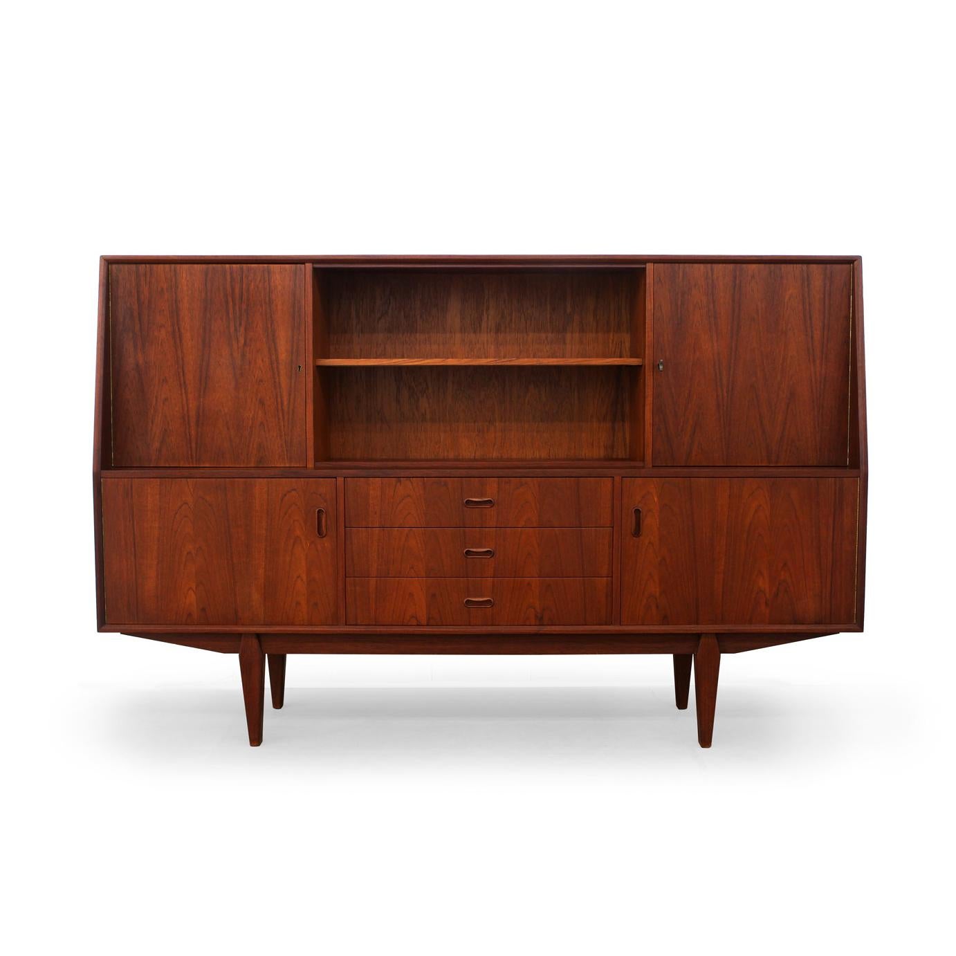 This teak Credenza features two upper locking cabinets with original key, an open shelf area with removable sliding glass doors, two lower cabinets and three drawers with dovetailed construction. This piece sits atop tapered legs and provides ample