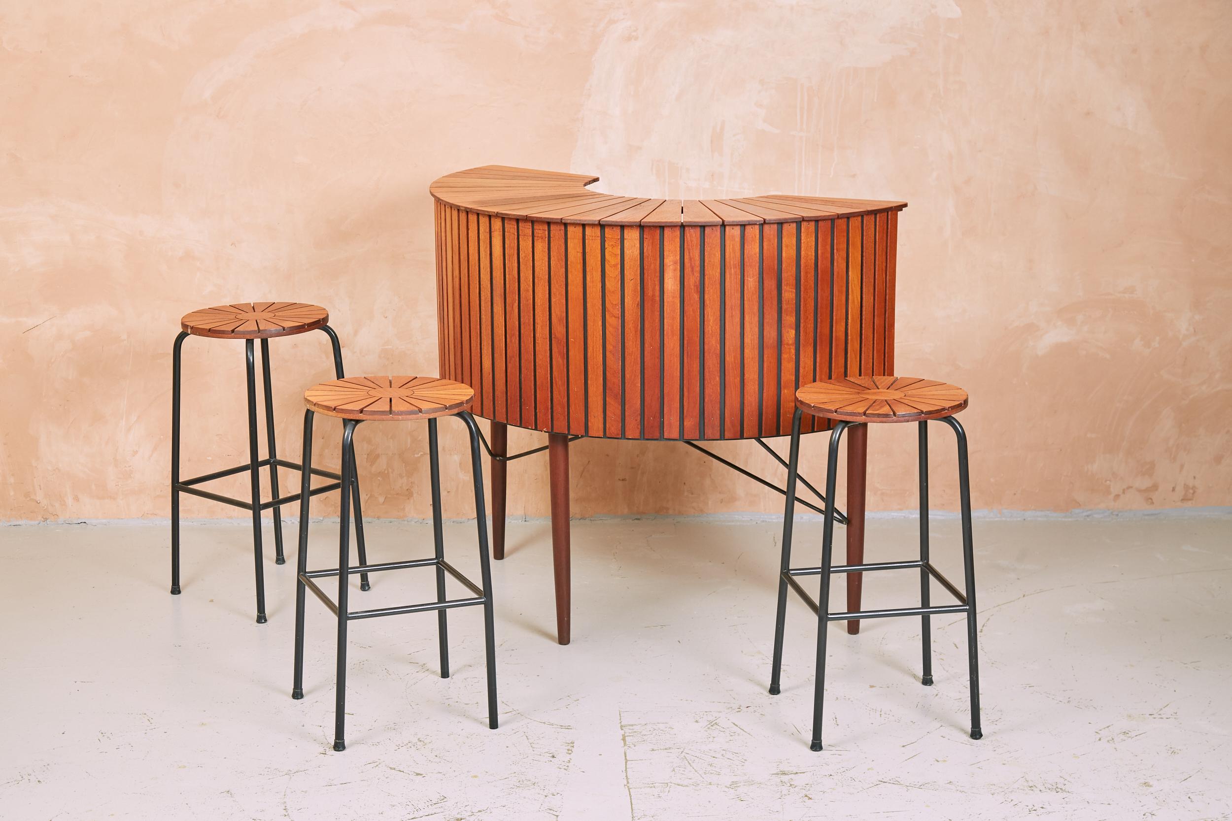 A rare and stylish 1960s home bar by Sika Møbler made in Denmark.
With a solid teak slatted bar, and matching stools in a sunburst pattern, this set is a real classic.
The demilune bar sits on solid teak legs, braced with black ironwork. There is